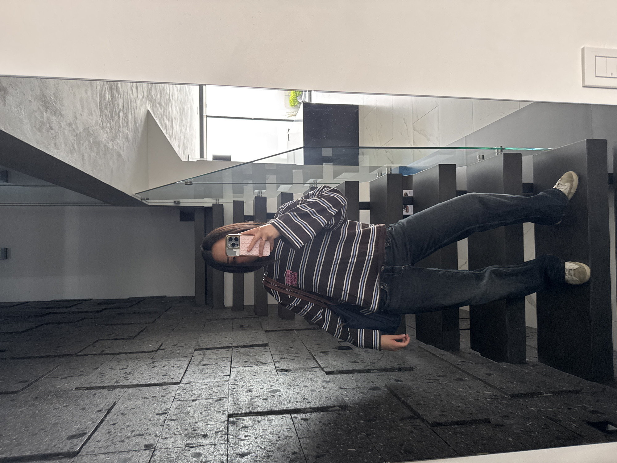 A person stands taking a selfie in a mirror in what appears to be an indoor space, possibly a building or a home, with a modern architectural design. The subject is wearing dark jeans, a horizontally striped long-sleeved shirt, and sneakers. The person holds a smartphone, the pink protective cover of which is visible, to take the photo.
The environment includes a staircase with a glass railing and dark steps, leading to an upper level. The floor is covered with dark, possibly textured, tiles. The lighting seems natural, coming through a window near the staircase.
The location is Puebla, Mexico.