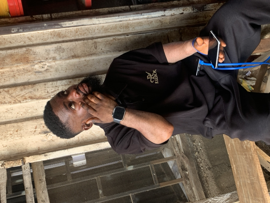 A male individual with a dark, short haircut and a full beard is depicted seated, looking upwards and to his right, with his left hand lightly touching his left cheek. He is wearing a black ribbed polo shirt with "REEBOK" and a wing-like logo embroidered in gold on the left chest, and black trousers. A dark-faced smartwatch is visible on his left wrist. In his right hand, he holds two smartphones, connected by a blue charging cable; one phone is plugged into the other, and the cable extends to his side. The individual exhibits what appears to be vitiligo on his right forearm and hand. He is seated on a rough wooden bench.
The background consists of various textures: to the left, a section of a dilapidated window or opening with a metal mesh screen and rough wooden framing. To the right, a corrugated metal wall with visible signs of wear and rust. Further right, a vertical red object and a yellow coiled item are partially visible. The ground area beneath the bench shows debris, including what appears to be crumpled paper or plastic.
The scene is located in the city of Okene, Nigeria.