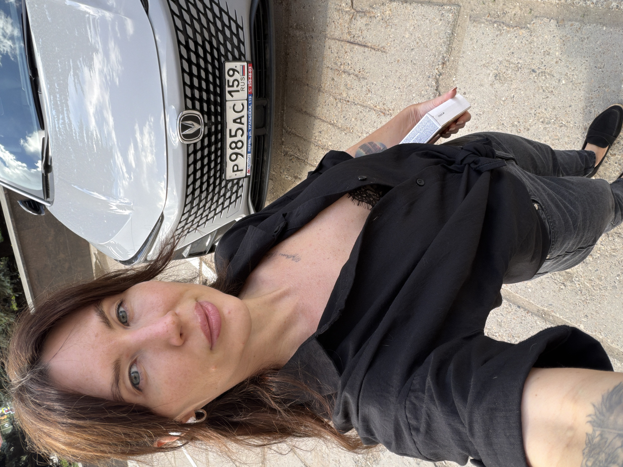 This media file is a portrait of a woman with long brown hair, green eyes, and light skin. She is wearing a black collared shirt, dark jeans, and black loafers. She is holding a small white box in her right hand. The background shows the front of a white car with a license plate that reads "P985AC 159". The setting appears to be outdoors on a paved surface, possibly a sidewalk or parking lot. The lighting suggests it is daytime, with clear skies visible on the car's hood, indicating sunny weather. The woman's expression is neutral, and she is looking towards the camera. Visible text on the license plate also includes "PERM", "URALSKAYA, 119", and "RUS". There is also some text on the white box she is holding, but it is not legible. The overall scene is casual, suggesting an everyday moment.