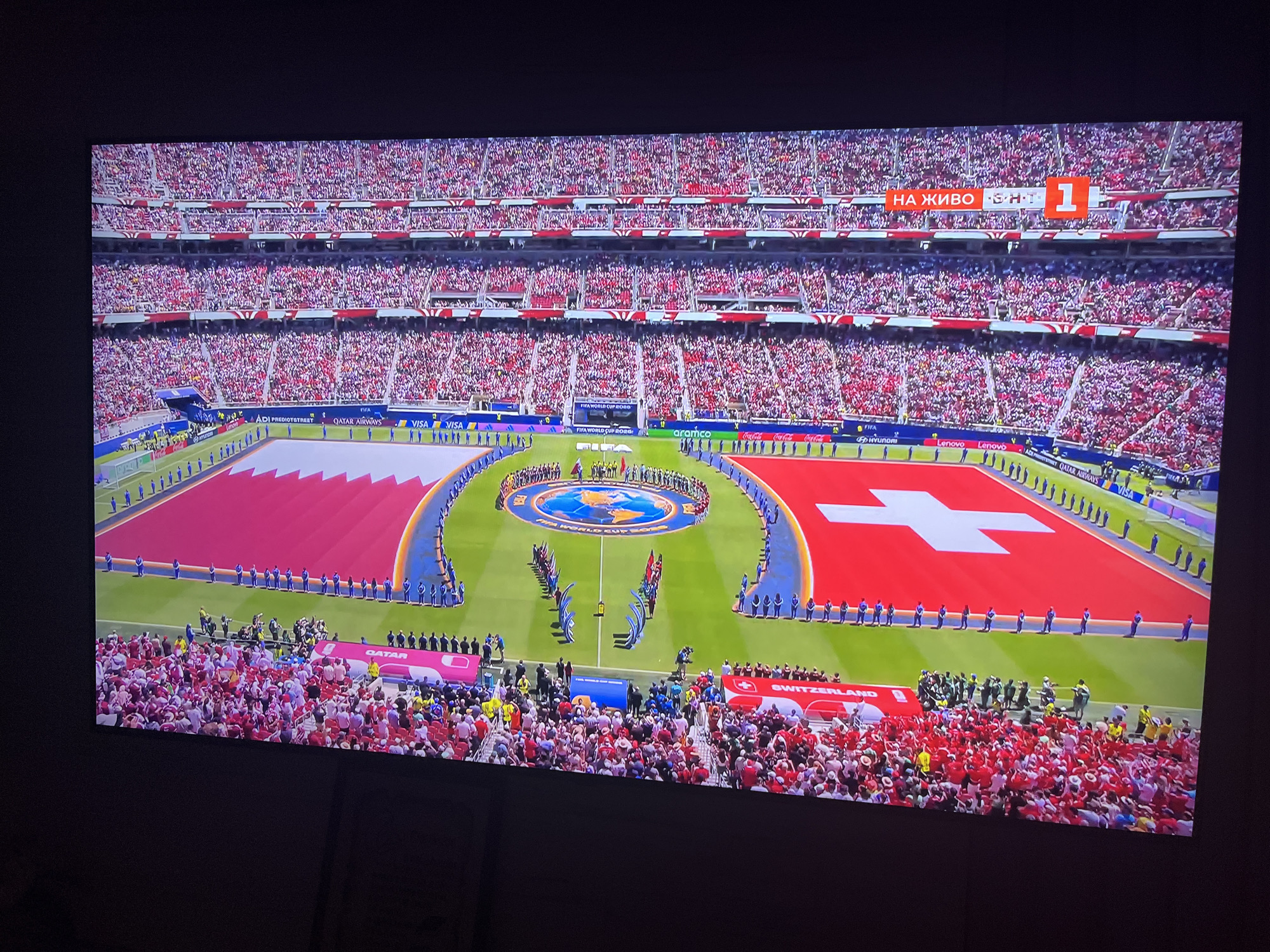 The image shows a large stadium filled with spectators, predominantly wearing red and white. The field displays two large flags: one is the flag of Qatar, and the other is the Swiss flag. Several individuals are standing on the field, some in blue uniforms, possibly players or officials. Along the edge of the field, there are banners and logos from sponsors such as FIFA, Qatar Airways, Visa, Lenovo, and Coca-Cola. The stadium itself appears to be a modern sports venue. The scene is captured on a television screen, indicated by the dark border around the image and slight glare. There is text displayed on the screen in orange boxes, with one reading "НА ЖИВО" (meaning "Live" in Bulgarian) and a score of "1-1". This suggests the event is a live broadcast of a sporting match, possibly a football game, which is being watched in Varna, Bulgaria.