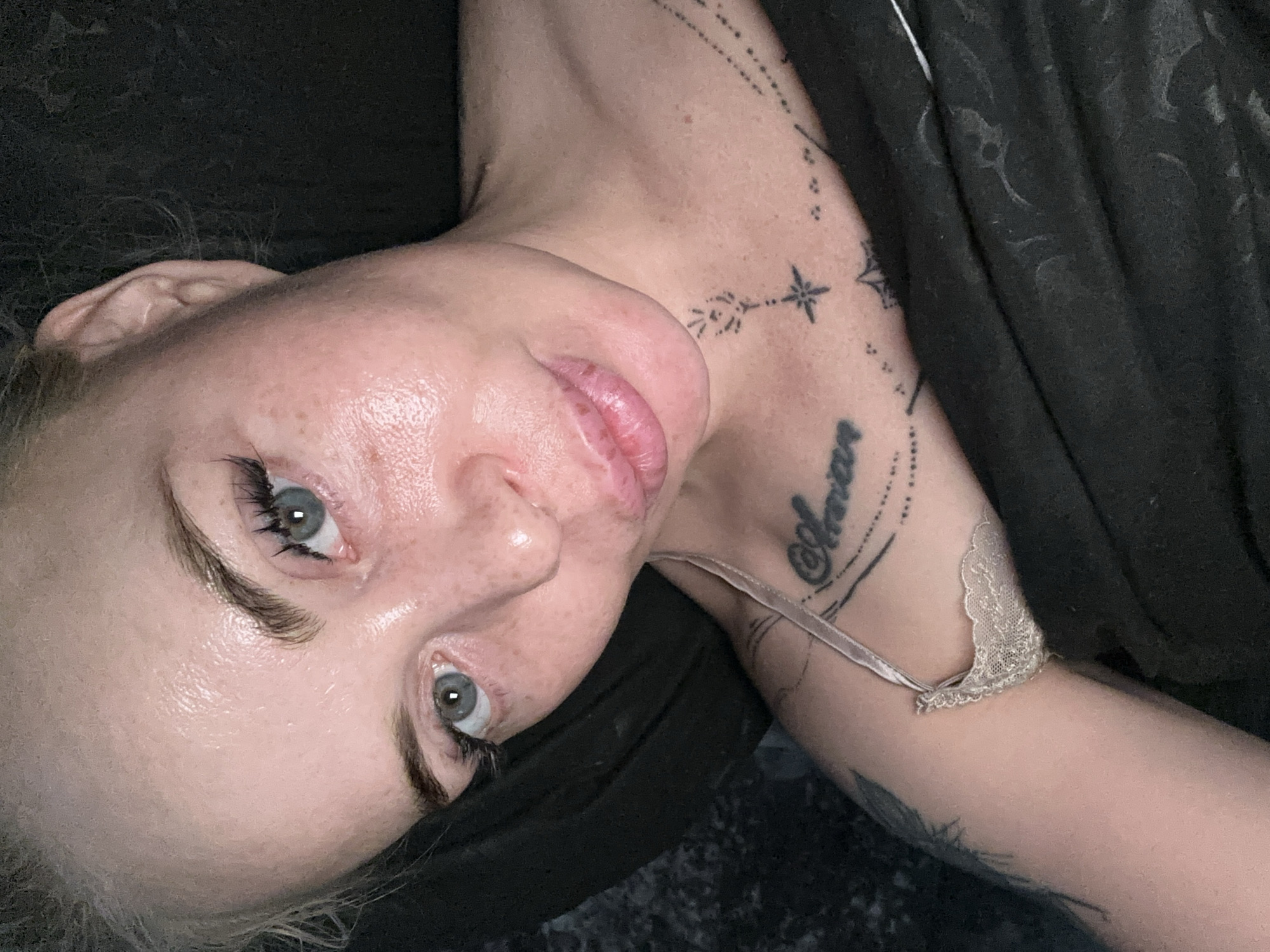 A close-up, overhead shot shows a fair-skinned woman with blonde hair pulled back. She has blue-green eyes, dark eyelashes, and light brown eyebrows. Her face is lightly powdered, and she has freckles. She is wearing a light-colored lace bralette and has tattoos on her chest and arm. She is lying down, and the background is dark. The scene appears to be indoors.