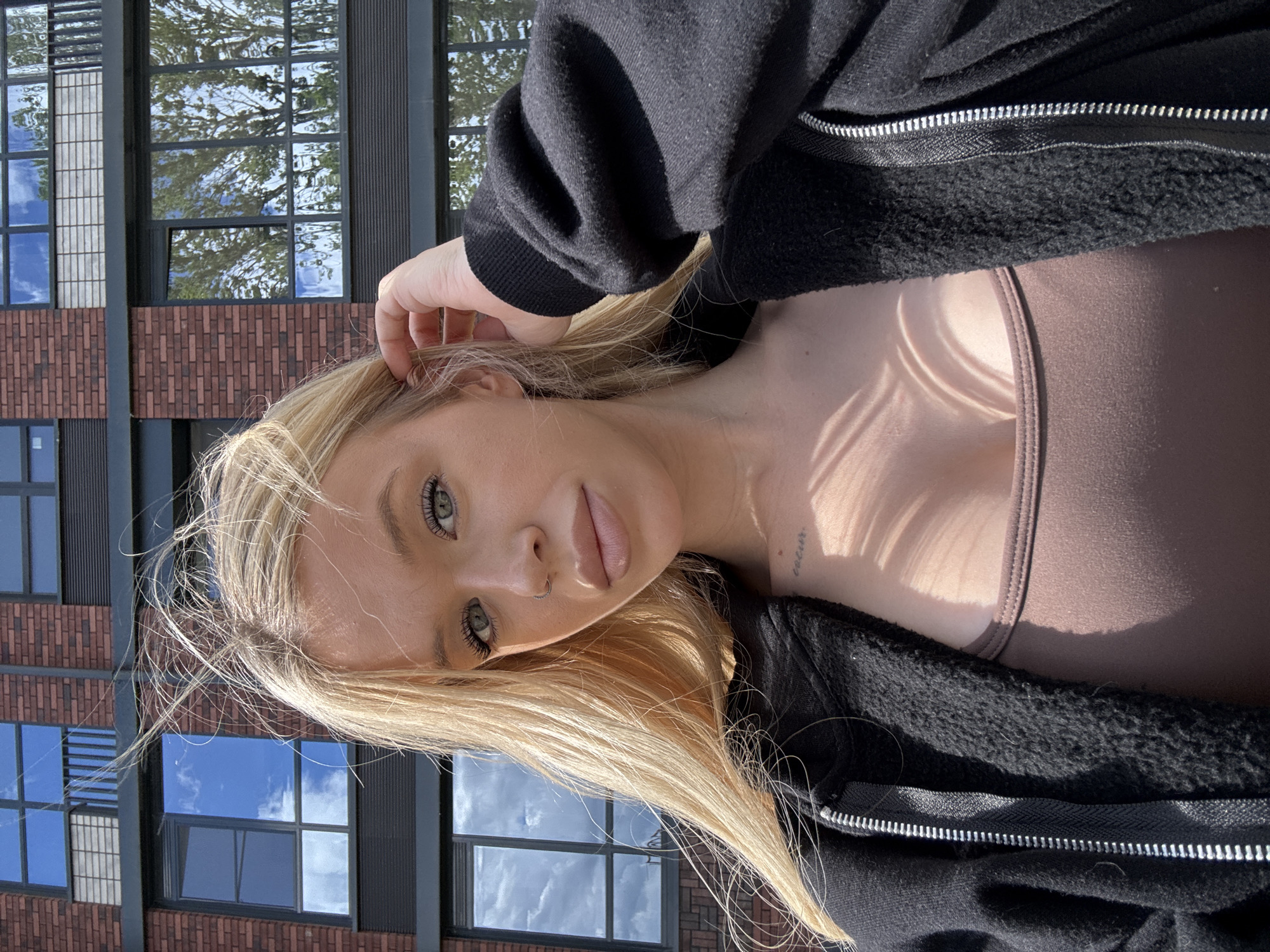 In Saint Petersburg, Russia, a young blonde woman with green eyes is captured in a selfie outdoors. She is wearing a brown tank top and a black zip-up hoodie. Her hair is windswept and falls across her face and shoulders. A small tattoo is visible on her chest. Behind her, a modern brick building with large windows reflects the blue sky and trees. The sun casts shadows from what appears to be foliage across her chest and hoodie. The woman is touching her hair with her right hand, and she is wearing a small hoop nose ring.