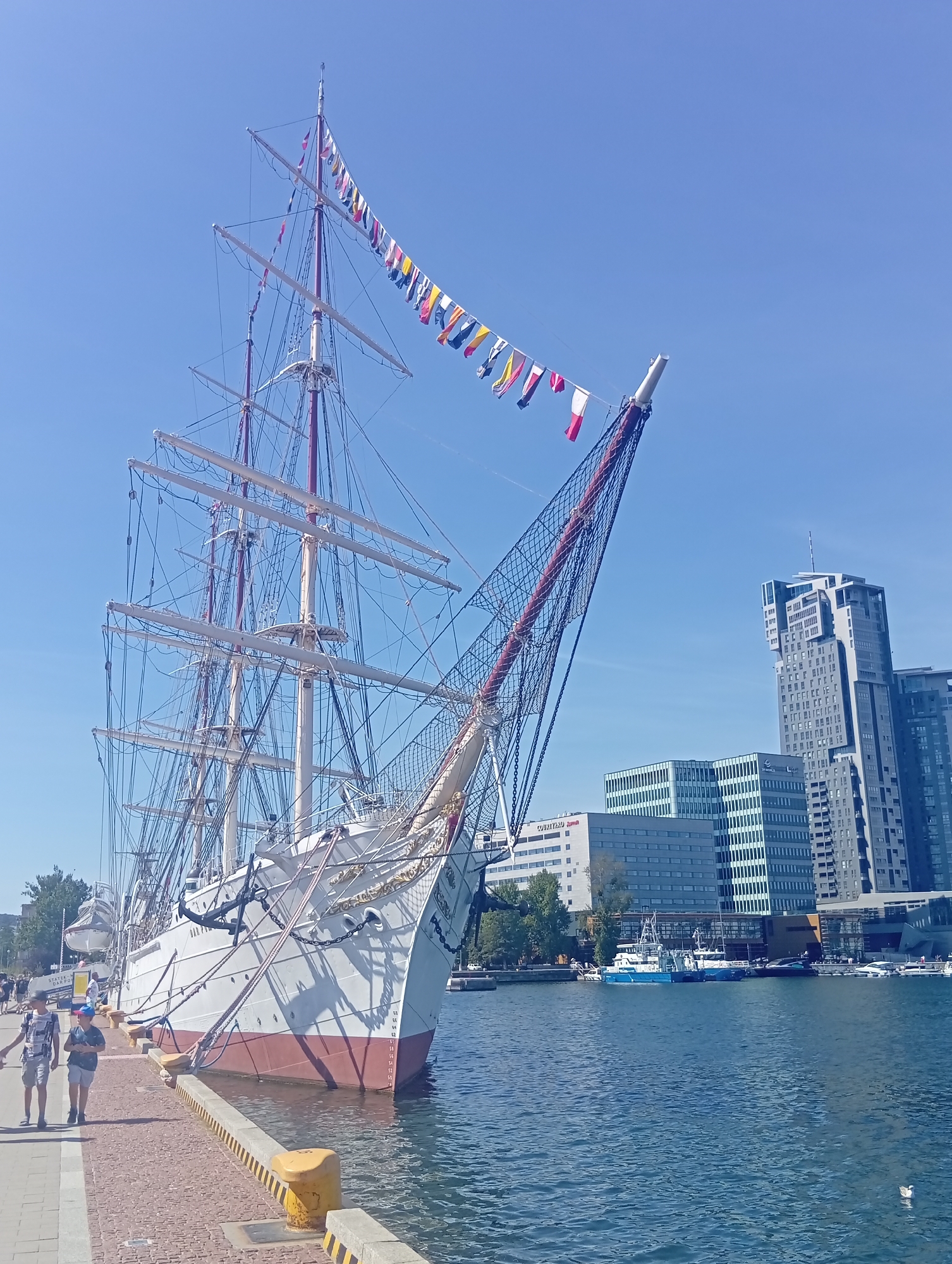 A large white sailing ship, the Dar Pomorza, is docked at a pier in Gdynia, Poland. The ship is adorned with numerous colorful maritime signal flags, strung between its masts and rigging against a clear blue sky. In the background, modern high-rise buildings dominate the city skyline. Two young boys are walking along the brick-paved pier in the foreground, to the left of the ship. The water is calm and reflects the blue sky.