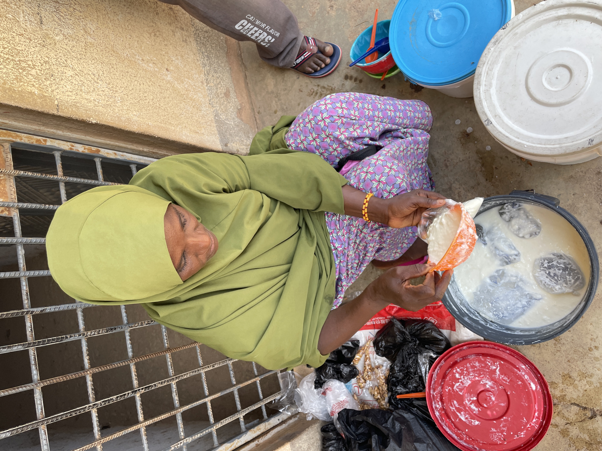 A woman in a green hijab and patterned dress squats next to a metal grate in Jos, Nigeria. She pours a white substance from an orange measuring cup into a bucket filled with a milky liquid and dark-colored circular objects. Nearby are several lidded buckets in red, white, and blue, along with black trash bags and walnuts. A man's legs and feet are visible at the top of the frame, wearing brown pants with "CHEERS!" printed on them and sandals. The ground is concrete. A wall is visible above the grate.
