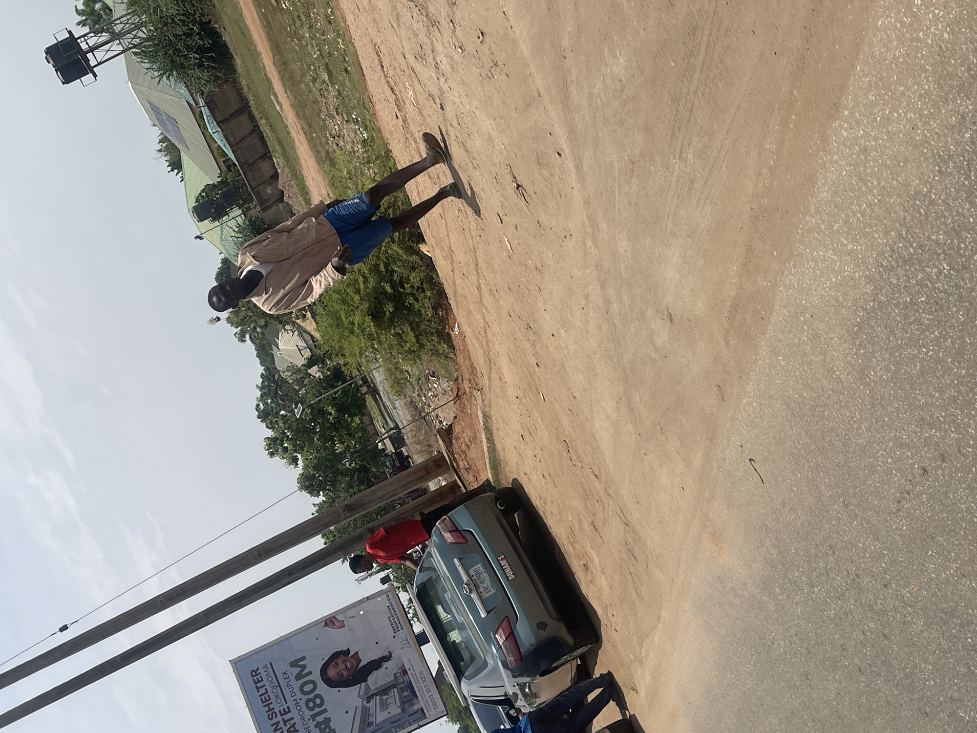 The image, rotated 90 degrees clockwise, captures an outdoor scene, likely a street or unpaved road in Galadimawa, Nigeria, under a clear, sunny sky during the daytime. The ground consists of a light brown dirt path bordering a grey, possibly paved, road surface.
In the mid-ground, a man wearing a light-colored long-sleeve top, blue shorts, and dark flip-flops stands on the dirt path, facing away from the camera, his shadow clearly visible. Adjacent to him, on the paved section, a light blue/teal sedan car is parked. The car's license plate reads "ABC-97580", and "DUNAM'S" is visible on its trunk.
Two other individuals are near the car: one in a red shirt is partially visible near the rear, appearing to lean into the car, and another, in blue clothing, is partially seen near the front, possibly squatting or bending. Power lines and utility poles diagonally cross the lower left portion of the frame.
A large billboard stands prominently near the car and poles, displaying real estate information: "AN SHELTER", "ATE LOKOGOMA", "BEDROOM DUPLEX", "#180M", a phone number "0803 312 3216", and "EZEKIEL PROPERTIES MANAGEMENT CO".
In the background, several buildings with light-colored, possibly metal, roofs are visible, some featuring solar panels. A tall metal structure, potentially a water tower or communication mast with two tanks, rises above the buildings, surrounded by green trees and bushes. Strong shadows indicate bright sunshine.