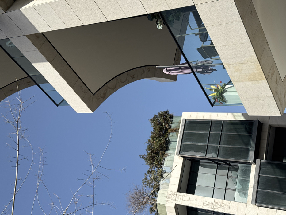 Captured from below, an architectural scene features beige, tiled buildings against a clear blue sky. A curved archway frames the upper portion, while a structure with dark-tinted windows stands diagonally. The windows reflect the surrounding architecture. Sparse branches reach into the sky from the lower left. A glass-enclosed space with plants and outdoor furniture is visible, further adding to the complex interplay of lines and reflections.