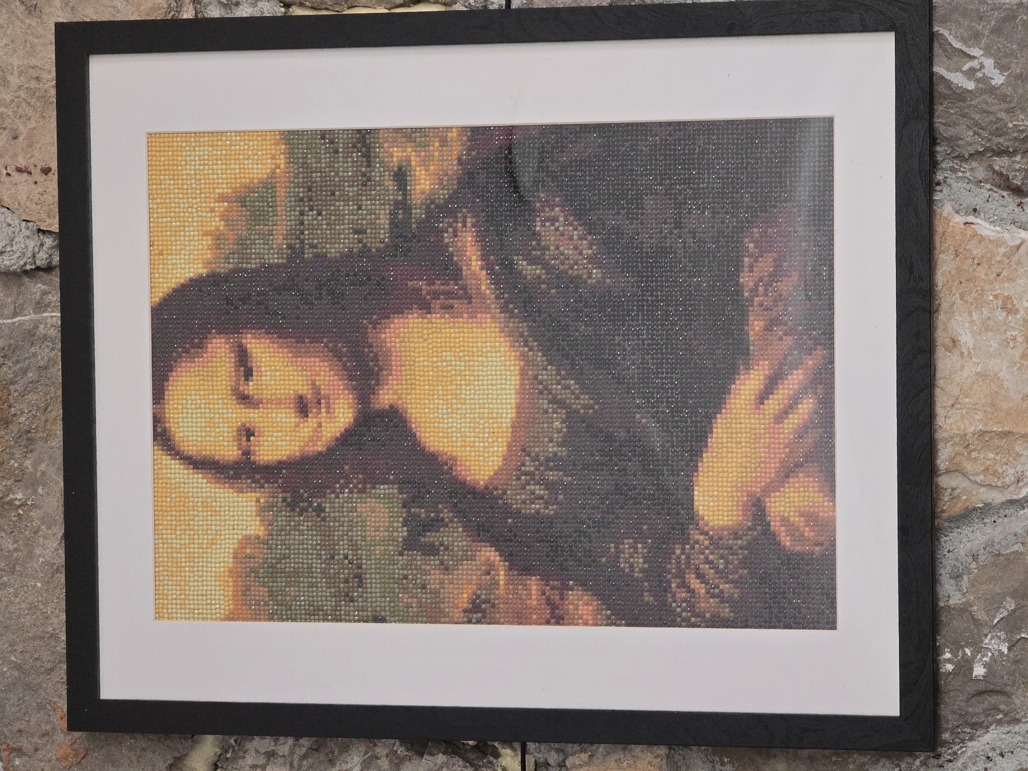 A framed diamond painting of the Mona Lisa is displayed against a stone wall in Podgorica, Montenegro. The painting, rendered in small, colored diamonds, depicts the iconic portrait with subtle variations in hue creating a textured, mosaic-like effect. The Mona Lisa is shown from the chest up, with her characteristic enigmatic smile and gaze. She wears dark clothing and her hands are clasped in front of her. Behind her, a landscape is visible, with muted tones suggesting trees and sky. The frame is black and simple, surrounding a white mat. The stone wall behind the artwork features a mix of brown, beige, and gray stones with rough textures, indicative of an outdoor or rustic interior setting.