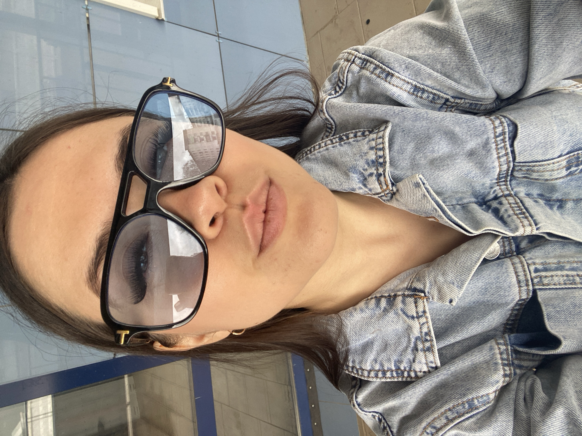 A woman is seen in a close-up selfie. She is wearing sunglasses and a denim jacket. The reflection in the sunglasses suggests an urban environment, possibly in the city of Chaikovsky, Russia. The background includes a blue and gray exterior, possibly a building facade.