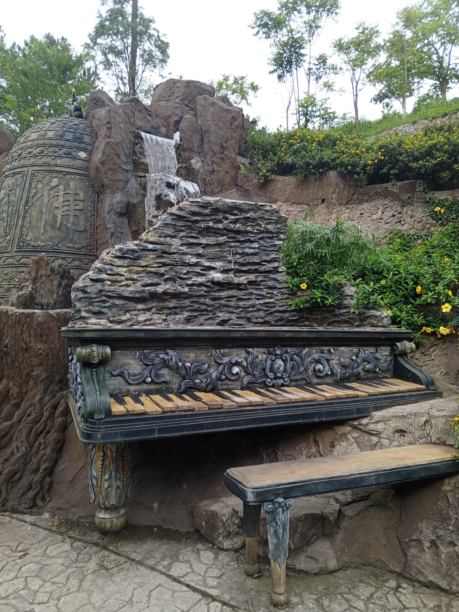 The image depicts a decorative piano-shaped structure with a bench in front of it, set against a rocky hillside. The piano structure is ornate with detailed carvings and has a keyboard made of alternating light and dark colored keys. Behind the piano, a small waterfall cascades down a rock formation. To the left of the waterfall and piano is a large, decorative bell, possibly a Buddhist bell, with intricate patterns and Asian characters inscribed on its surface. Lush green foliage, including bushes with small yellow flowers, covers parts of the hillside. The ground in front of the structure is paved with irregular flagstones. The scene appears to be an outdoor art installation or a garden feature, possibly located in a tourist area. The presence of the bell and its inscriptions suggest a possible connection to Buddhist culture, which is prevalent in Vietnam. The environment is naturalistic with rocks and greenery, indicating an outdoor setting. The overall impression is one of artistic landscaping and cultural representation. This decorative piano installation is located at the Datanla Waterfall in Da Lat, Vietnam.
