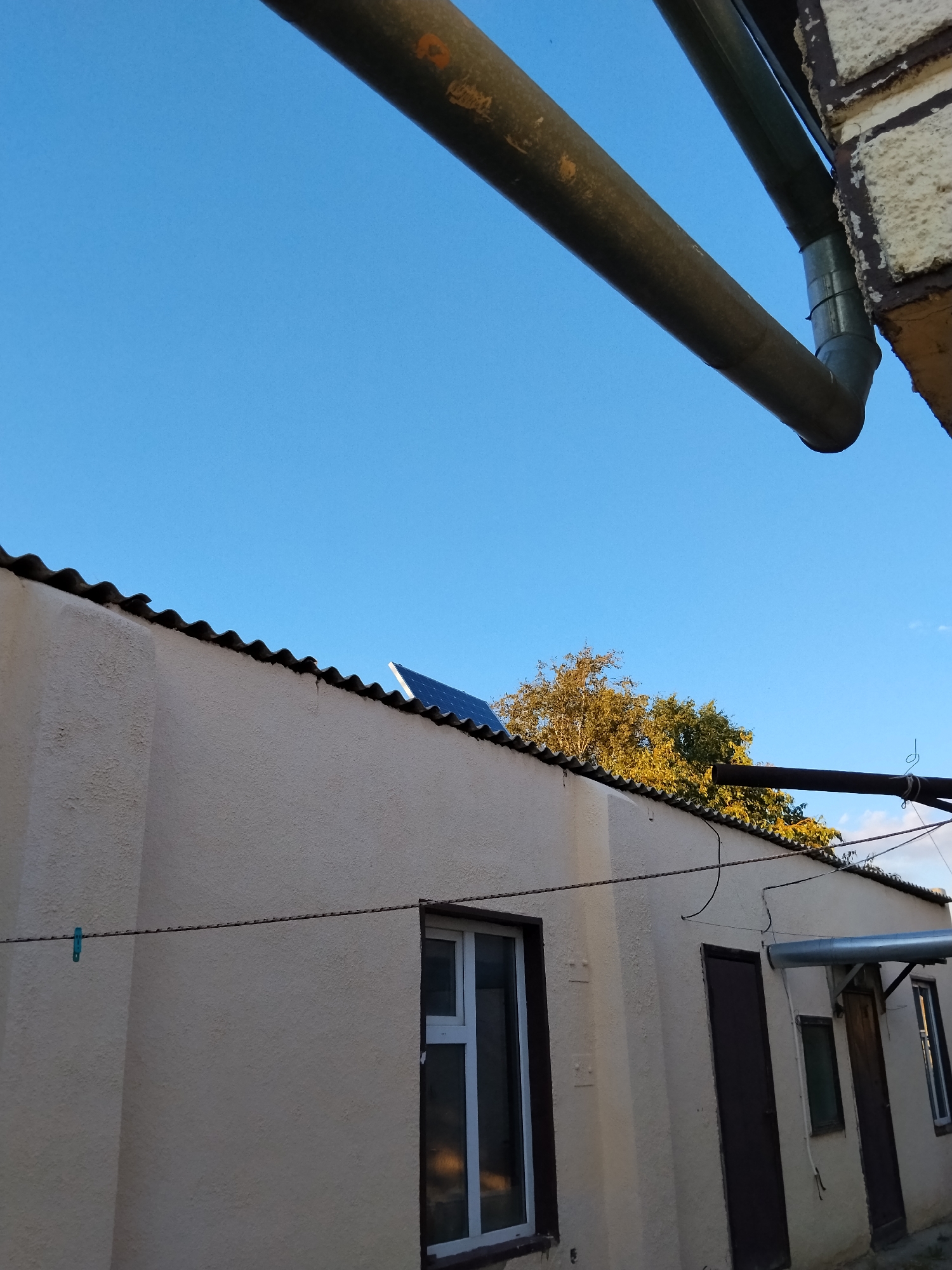 The image shows the side of a light-colored building under a bright blue sky. A corrugated roof extends over the building. A large, dark pipe runs diagonally across the top of the image, supported by a brick structure on the right. A smaller pipe, possibly a downspout, connects to the larger pipe and curves downward. A solar panel is visible on the edge of the roof. Several windows and doors are present on the building's wall. A clothesline with a blue clothespin stretches across the wall. Trees with yellow and green foliage are visible behind the building. The scene appears to be an outdoor setting in Kokshetau, Kazakhstan, likely in a residential or utility area. There are no people or animals visible in the image.