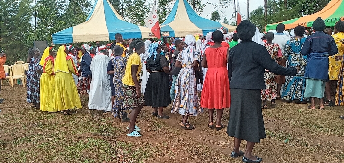 A large gathering of people, primarily women, are present in an outdoor setting, likely a community event, located in Lwandeti, Kenya, at the coordinates provided. The scene unfolds beneath the shade of a large tent with a blue and white striped canopy. The people are dressed in colorful clothing, many in bright yellow and red dresses. Some are wearing traditional African patterns. A few men are also present, standing in the background and participating in the gathering. The atmosphere is lively and celebratory. The women appear to be engaged in social interactions, possibly dancing or singing, while the men are observing. The ground is covered in a thick layer of grass, typical of an outdoor setting. The area is surrounded by trees and greenery, creating a natural and vibrant environment. It is a joyous occasion with a strong sense of community engagement.