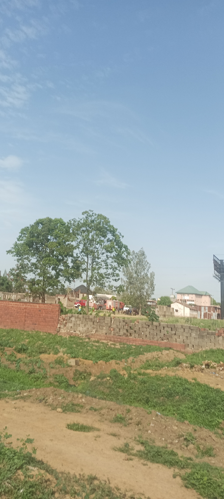 The image shows an outdoor scene in Jos, Nigeria, during daytime. The sky is blue with a few clouds. In the foreground, there is a dirt and grassy area. Behind that, a brick wall composed of red bricks and cinder blocks runs across the midground. Behind the wall are several vehicles, including fire trucks, and a small group of people. Two large green trees are near the left side of the image. Other buildings and structures are visible in the background, including a pink building and a billboard.
