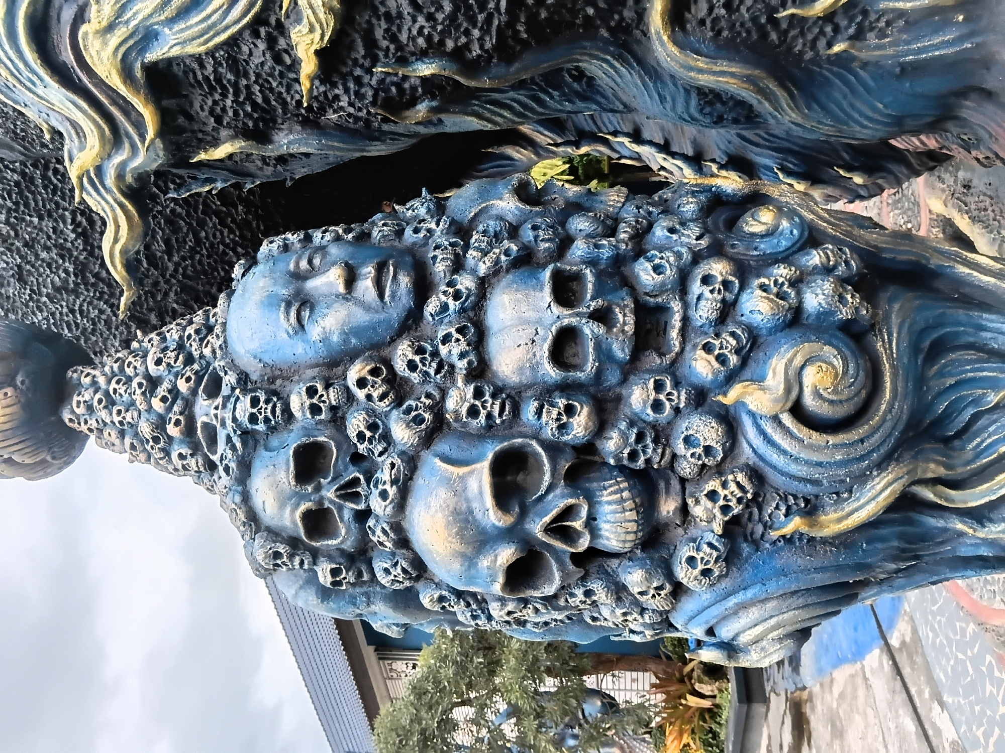 The image shows a close-up of a striking, blue and gold statue composed of numerous skulls and at least one human-like face, arranged in a dense, layered manner. The statue is intricately detailed, with a textured, almost rough finish on some areas and smoother, more polished sections on others. The skulls vary in size, with some significantly larger than others, creating a sense of depth and mass. The human face is positioned prominently among the skulls, with its eyes closed and an enigmatic expression. The statue is adorned with swirling, flame-like elements in gold and blue, adding a dynamic and potentially menacing quality.
Adjacent to this sculpture is a large, dark, textured structure, possibly another part of the same artistic installation, also featuring blue and gold accents in a flowing, wave-like pattern. The background reveals elements of an outdoor setting. To the left, a building with a dark tiled roof and light-colored walls is visible, along with some greenery, including a tree with sparse foliage and some ornamental plants. The ground appears to be paved with tiles, some of which are dark and some light, creating a mosaic-like pattern. The sky above is overcast with a cloudy, grey hue.
This type of artwork is characteristic of the unique and often macabre sculptures found at the "Baan Dam Museum" (Black House Museum) or the "Wat Rong Khun" (White Temple) and its associated grounds, which are prominent art installations located within or in the immediate vicinity of Chiang Rai City Municipality, Thailand. While the specific location within Chiang Rai cannot be definitively pinpointed solely from the image without further context, the style strongly suggests it is part of one of these renowned artistic complexes.