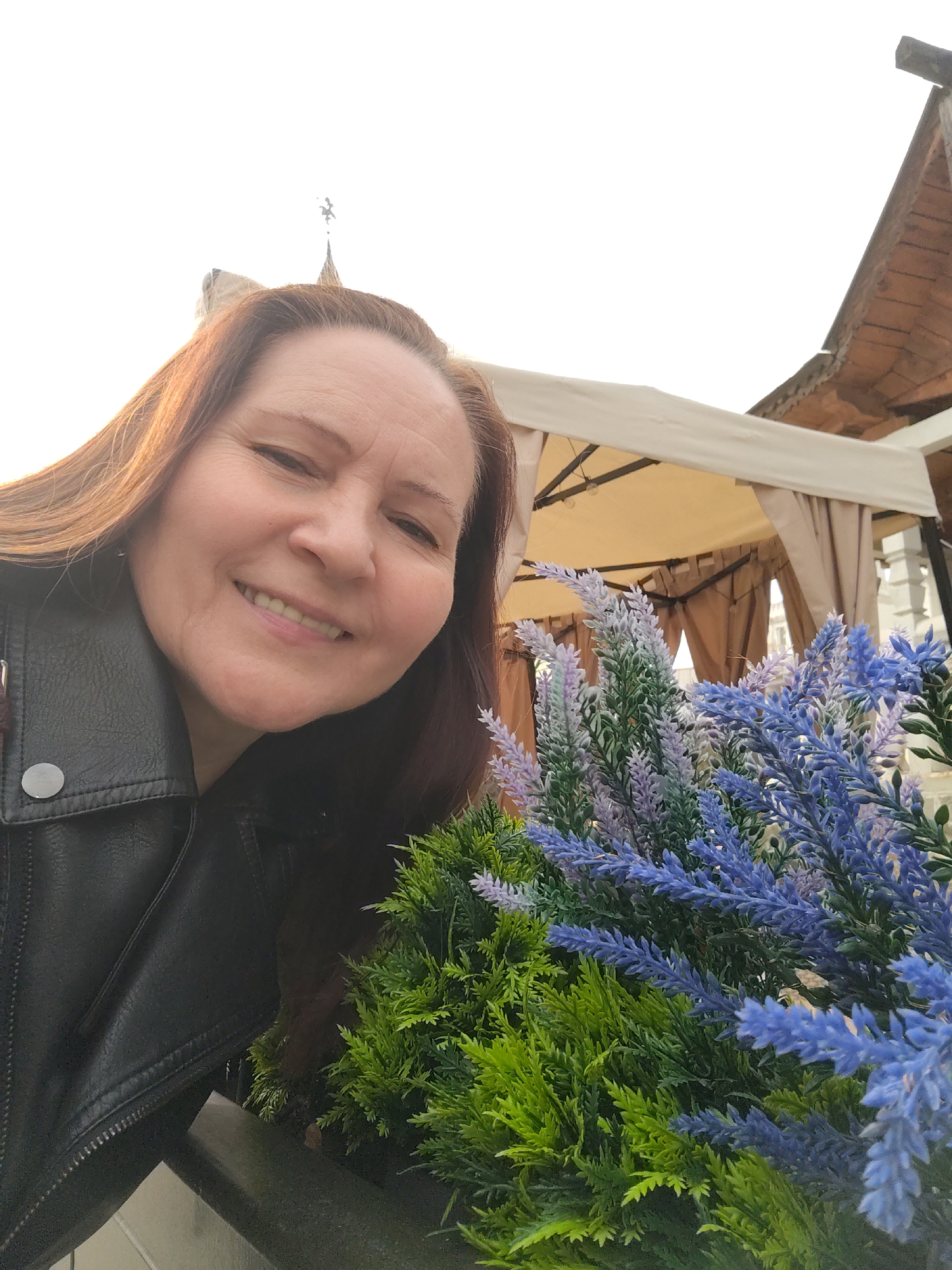 A smiling middle-aged woman with long, reddish-brown hair is captured in a close-up outdoor selfie. She looks directly at the camera with a wide, friendly smile, her head slightly tilted. She is wearing a black leather jacket with silver button details on the collar.
In the foreground and mid-ground, to the right and bottom of the frame, are decorative artificial plants. These consist of vibrant blue and purple lavender-like sprigs intermingled with bushy green foliage.
The background reveals a bright, clear sky on the left side. Towards the right, an outdoor structure is visible, featuring a light-colored, possibly beige or cream, fabric canopy or gazebo. Beyond this canopy, parts of a darker, possibly tiled or wooden, roof structure can be seen. In the upper center, a faint silhouette of a small spire or decorative architectural element is visible against the bright sky, hinting at a building in the distance.
The scene is well-lit, indicating it is daytime. The woman's expression conveys happiness and warmth. The setting appears to be an outdoor cafe, restaurant area, or a public garden. Based on the provided context, the image was taken in Moscow, Russia. No visible text is present in the image.