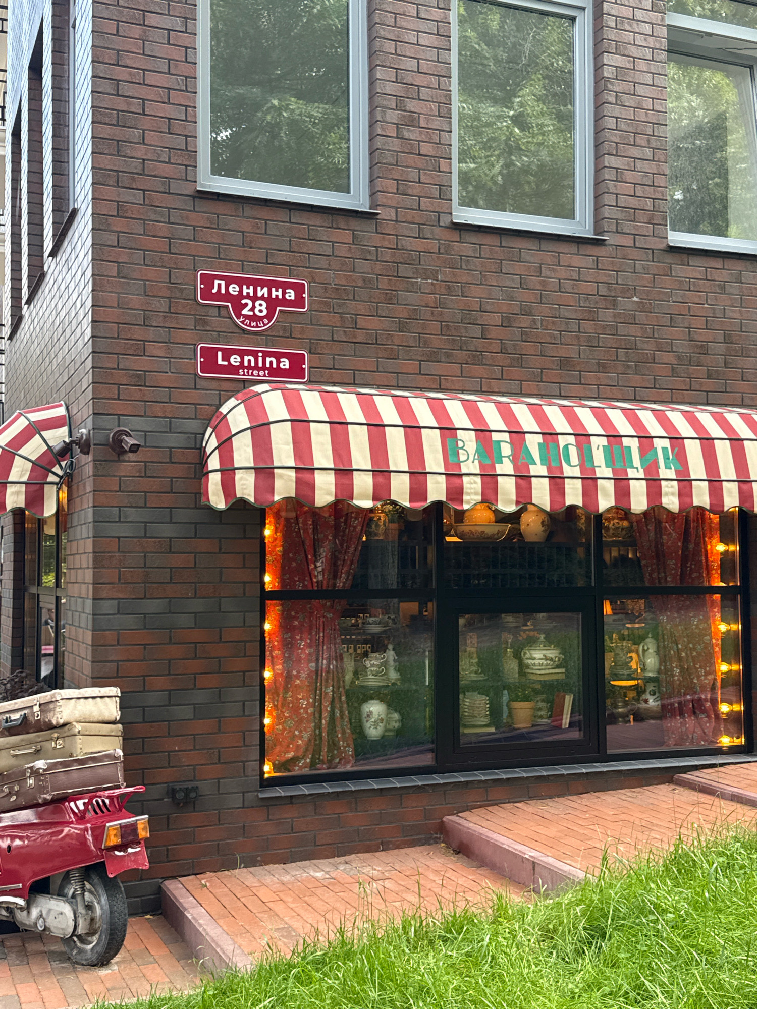 A red motor scooter is parked on a brick pathway in front of a brick building. The motor scooter is loaded with three vintage suitcases. Above the scooter, the building has a large window with a red and white striped awning. The awning has the Cyrillic text "БАРАНОЧНИК" written on it in green letters. Two street signs are visible on the brick wall. The top sign reads "Ленина 28 улица" and the bottom sign reads "Lenina street". Trees are reflected in the building's windows. The area is overgrown with green grass.