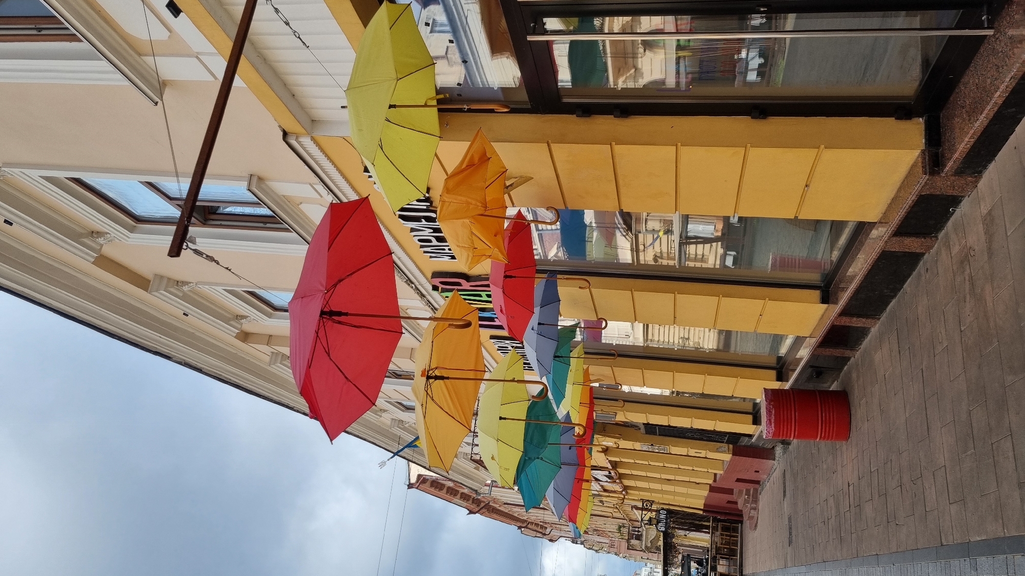 The image depicts an outdoor scene on a cloudy day, likely on a street in Chernivtsi, Ukraine. The foreground shows a pedestrian walkway paved with rectangular stone tiles. To the left, a red cylindrical barrel is placed on the sidewalk.
The midground is dominated by the facade of a building painted a bright yellow. Numerous colorful umbrellas—red, yellow, orange, teal, blue, and dark gray—are suspended from wires across the street, creating an overhead canopy. These umbrellas are open and hang at varying heights. The building features large, reflective windows and ornate architectural details on its upper level, including cornices and decorative elements. A sign with Ukrainian text, possibly a store name, is visible above the windows.
The background shows the continuation of the building and street, with some architectural elements suggesting older construction. The sky is overcast, contributing to the diffused lighting of the scene. There are no visible people or animals in the image. The overall impression is an artistic street decoration enhancing an urban environment.