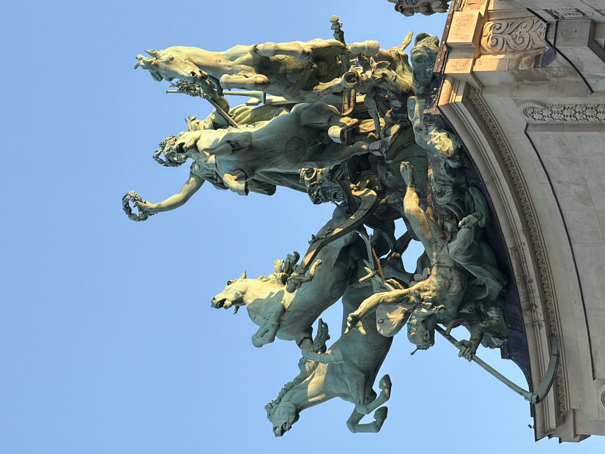 The image captures a close-up, low-angle view of a sculptural group attached to a monumental arch. The sculpture features multiple horses pulling chariots. Several figures are depicted, including warriors, a figure holding a wreath, and a figure wielding a scythe. The sculpture is a pale, weathered green color, typical of bronze. The architectural elements of the arch are pale stone with detailed carvings. The background is a clear, pale blue sky. This sculpture is located on the Arc de Triomphe in Paris, France.