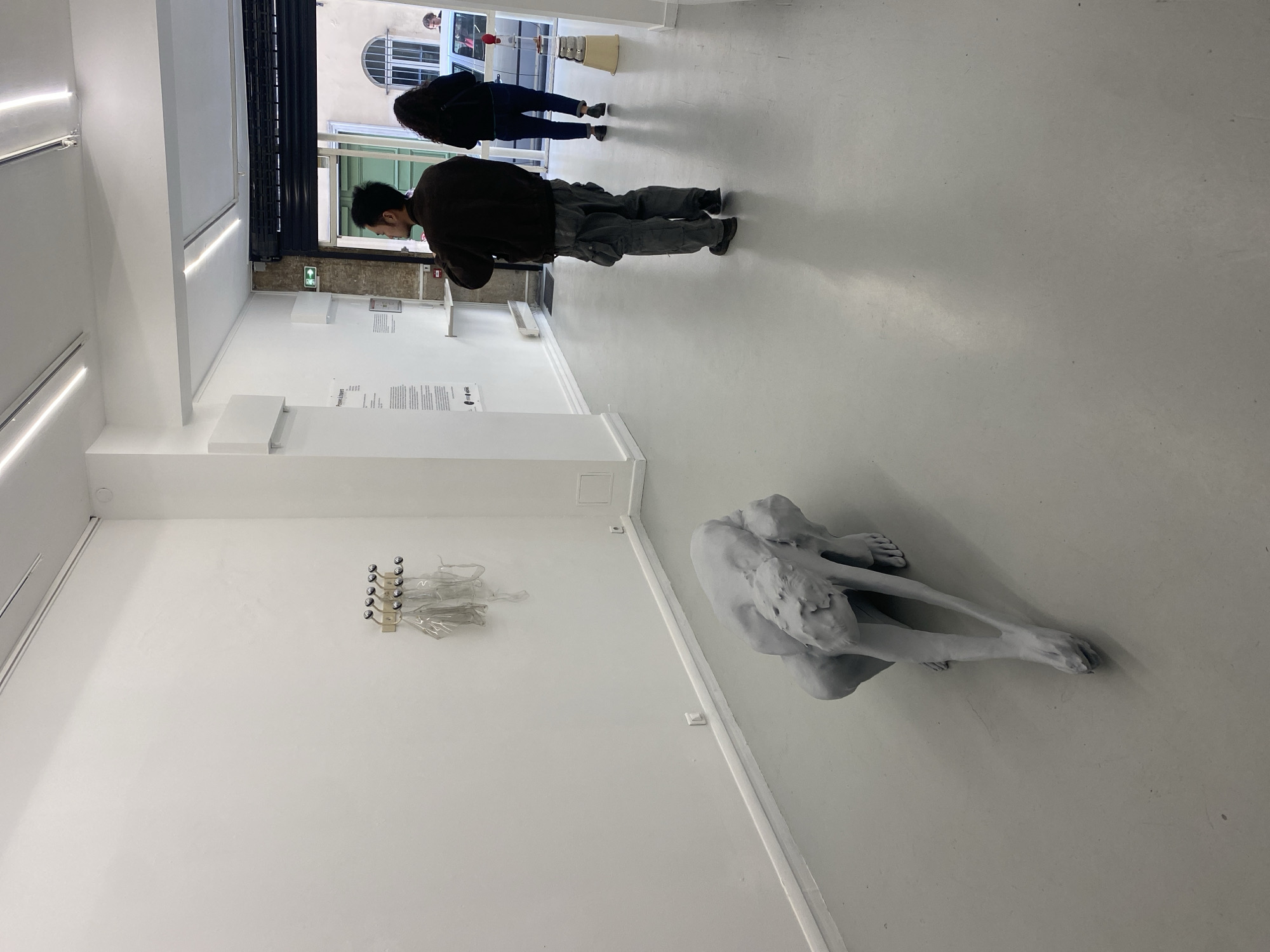 The image depicts the interior of a minimalist art gallery, characterized by stark white walls and a light-grey floor. The space is well-lit by artificial strip lighting along the walls and ceiling, as well as natural light filtering in from an opening at the far end.
In the foreground, to the right, a large, light-grey sculpture of a human figure is positioned on the floor. The figure appears to be in a fetal or curled-up pose, conveying a sense of introspection or vulnerability. On the left wall, a smaller art piece consists of a metallic rack with several round knobs, from which translucent, clear plastic or fabric elements are draped. Further along this wall, multiple white plaques with black text are displayed, providing information about the exhibition or artwork; one visible title is "Pierre Lescure."
Two people are seen from behind in the background, walking away from the camera towards the gallery's exit. A man wearing a dark brown jacket and grey cargo pants is in the mid-ground, while a woman in a dark jacket and jeans is further back. They appear to be observing the gallery space or heading towards the exit. Through the doorway at the back, an arched window and a glimpse of an exterior building are visible, hinting at an urban setting, consistent with Paris, France. A black roller shutter is partially lowered above the doorway.
The scene conveys a calm, contemplative atmosphere typical of an art exhibition. The time of day appears to be daytime.