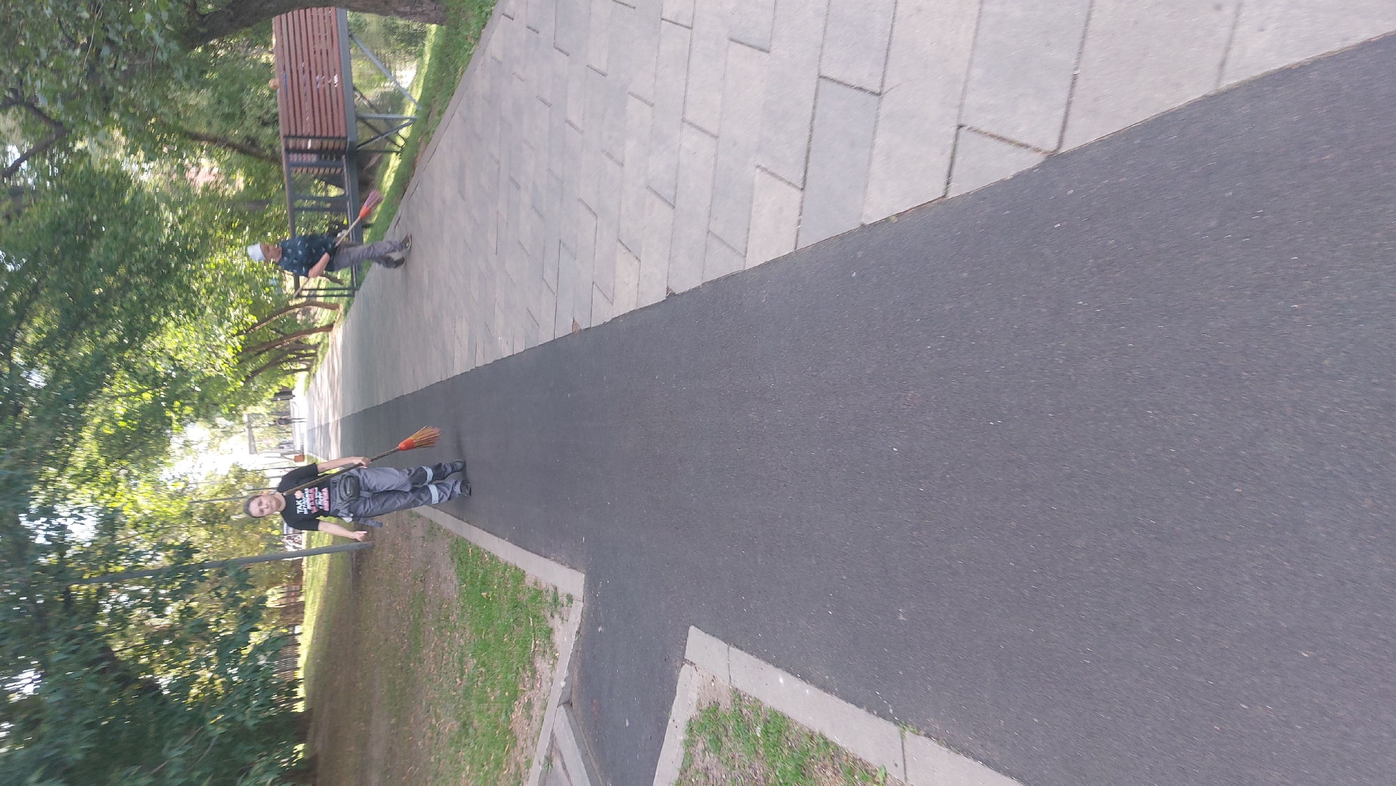 A woman in gray overalls and a black t-shirt sweeps a pathway with a broom. She is on a dark asphalt path that runs alongside a lighter-colored paved area. Trees line the path, and there is a wooden bench in the background. Another person in a blue shirt and white hat also sweeps a path further down. The scene appears to be a park in Moscow, Russia.