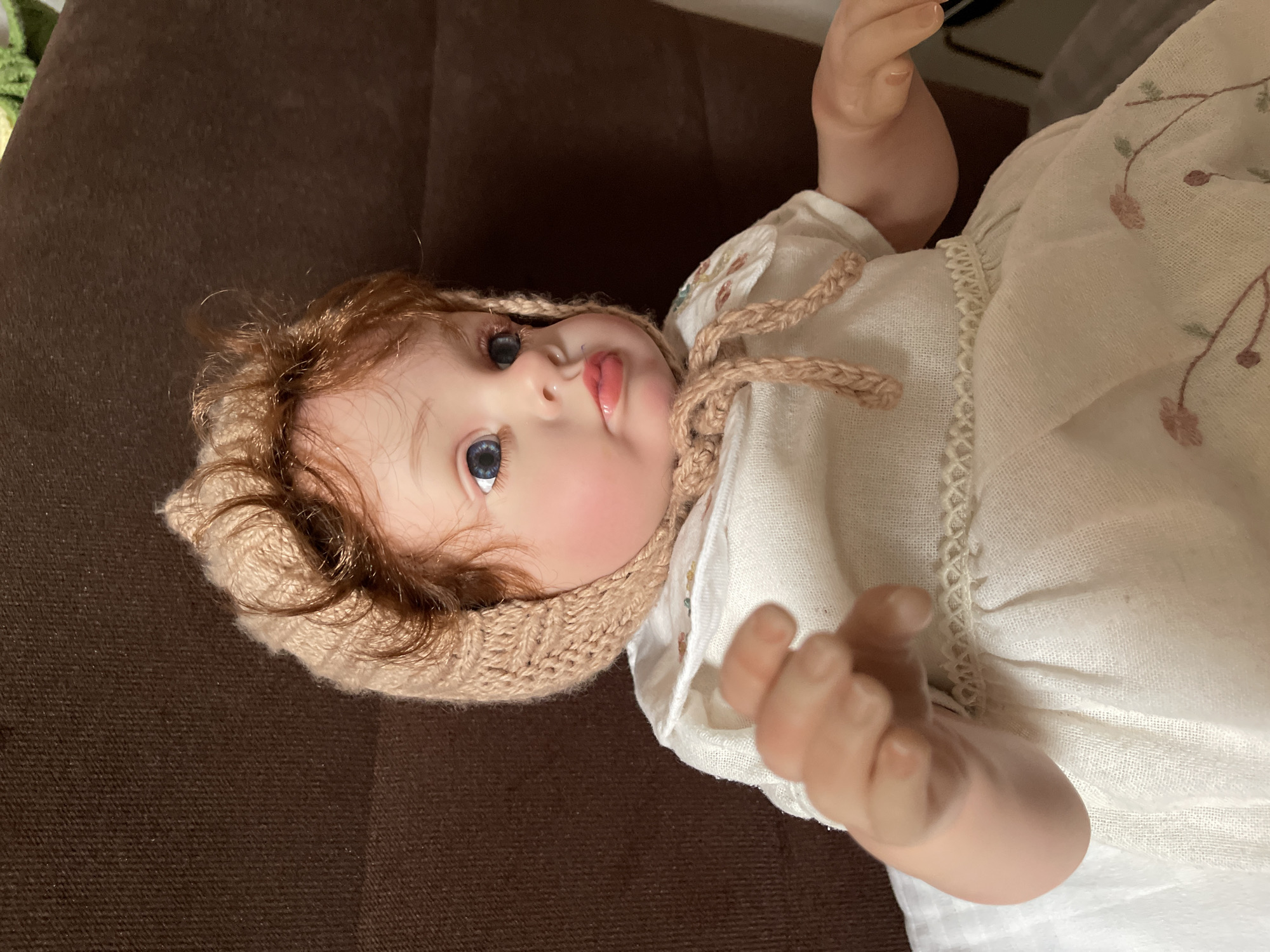 A realistic baby doll with blue eyes, rosy cheeks, and auburn hair is pictured indoors. The doll wears a knitted bonnet, a white dress with floral embroidery, and is seated against a brown textured background, likely a couch or chair. The doll's left hand is positioned in the foreground, appearing to reach forward. This image was taken in Zelenogradsk, Russia.