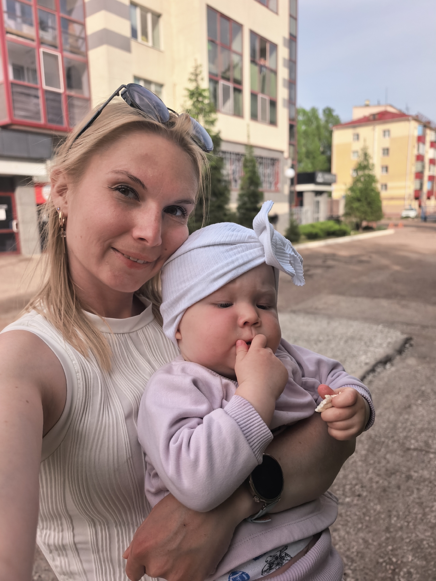 A fair-skinned woman with blonde hair and sunglasses perched on her head holds a fair-skinned baby wearing a white headwrap and a lavender-colored outfit. The baby is putting fingers in their mouth and holding a piece of food in their other hand. The woman is wearing a sleeveless white top. The background includes multistory buildings in Sterlitamak, Russia. The ground is concrete.