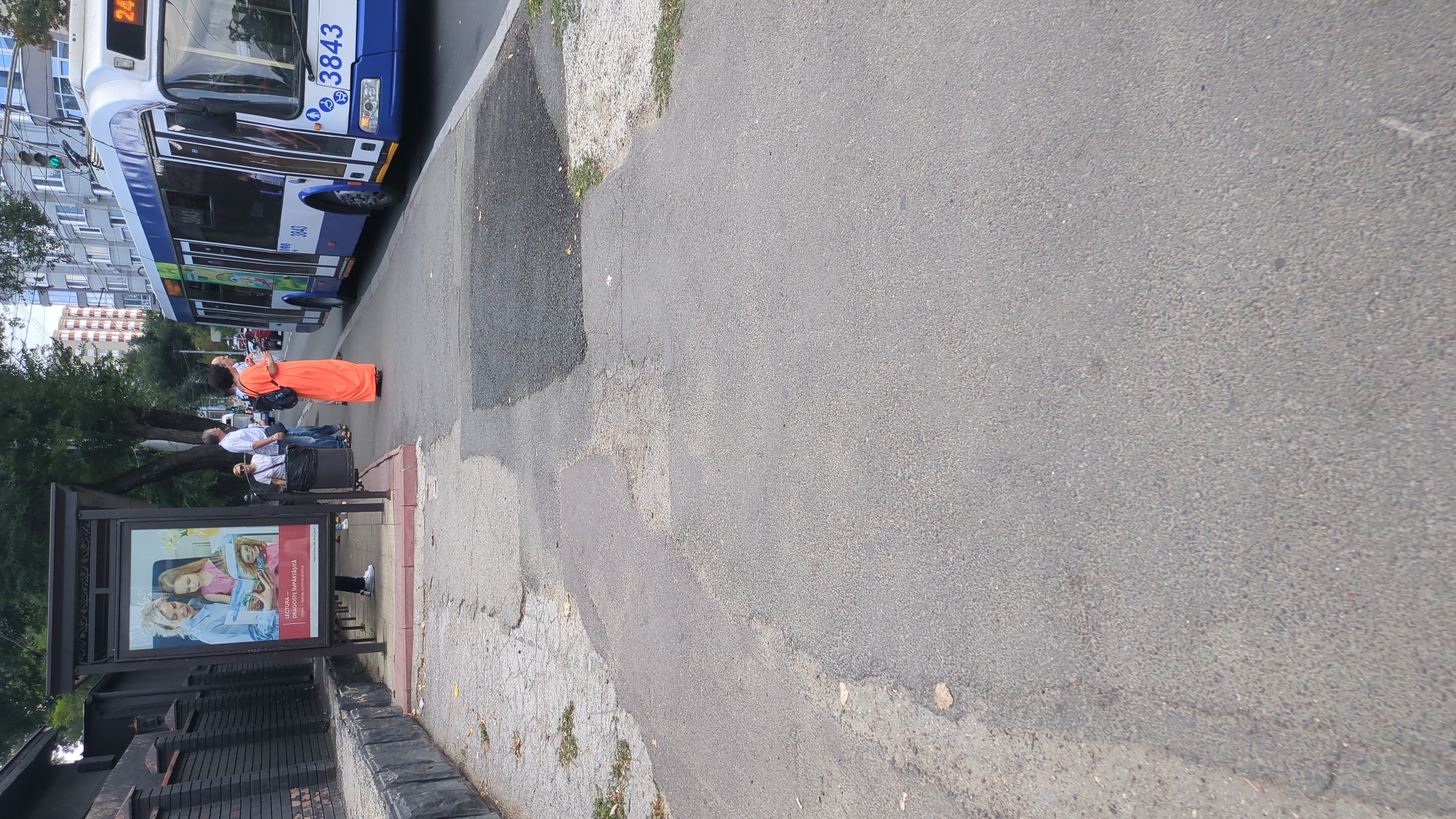 The image shows a street scene in Chișinău, Moldova. In the foreground, a paved sidewalk is visible, with some patchy repairs and fallen leaves. To the left, a dark brick wall lines the sidewalk. A bus stop shelter is present, featuring an advertisement depicting two women reading a book, with the text "LECTURA ÎMPĂRTĂȘITĂ" (Shared Reading). Several people are gathered at the bus stop. A woman in an orange dress stands facing the street, while a man in a white shirt and jeans and another man in a light blue shirt and dark pants are also present. A white trolleybus with the number 3843 is parked alongside the curb, its doors open. The background shows buildings and trees, indicating an urban environment.