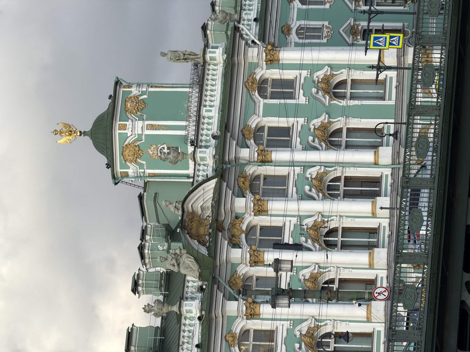 This image shows a close-up, eye-level view of the Winter Palace in Saint Petersburg, Russia, on a cloudy day. The palace is a large, green and white building with ornate gold embellishments. The architecture is neoclassical, with prominent columns, arched windows, and statues of figures. On the roof, there are several chimneys, sculptures of mythical creatures and human figures, and a golden double-headed eagle crowning a green dome. In the foreground, a metal railing of a bridge crosses the bottom of the frame, with cars visible on the road beyond. Traffic signs, including pedestrian crossing signs and a no-pedestrian sign, are also visible. Electrical wires crisscross the scene, partially obscuring the building. The sky is overcast, with a muted grey tone. The scene suggests a busy urban environment, with the grandeur of the palace contrasted by the functional elements of modern infrastructure.