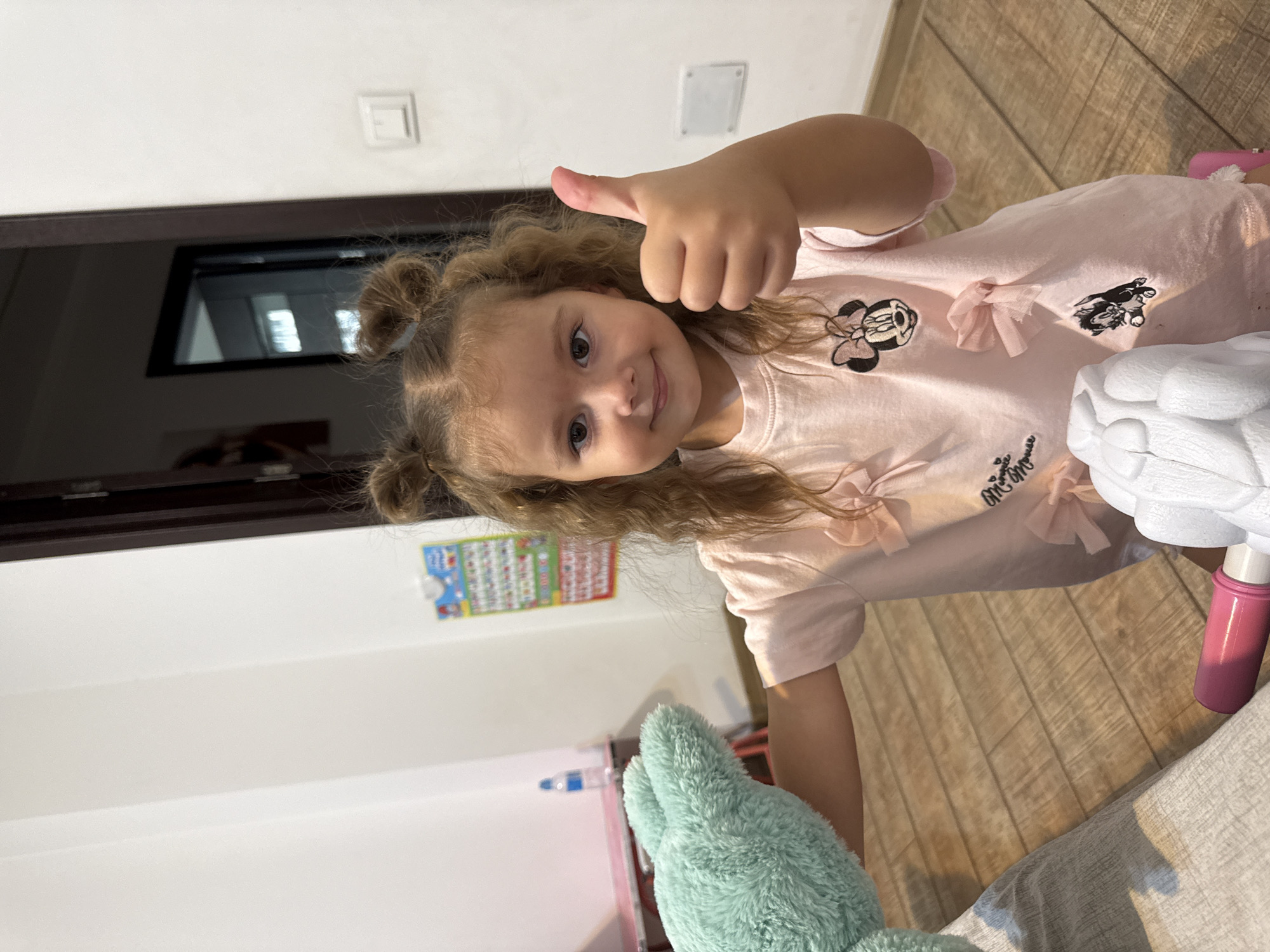 A young girl with curly blonde hair styled in two buns is standing in what appears to be a room in Chișinău, Moldova. She is wearing a pink t-shirt with Minnie Mouse and other cartoon characters printed on it, adorned with small bows. She is holding a light green plush toy in her left arm and giving a thumbs-up with her right hand. The room has white walls, a wooden floor, and a dark wooden doorframe visible in the background. There is a light switch on the wall and a colorful chart on the wall behind the girl. A pink object, possibly part of a toy, is visible on the floor in the foreground.