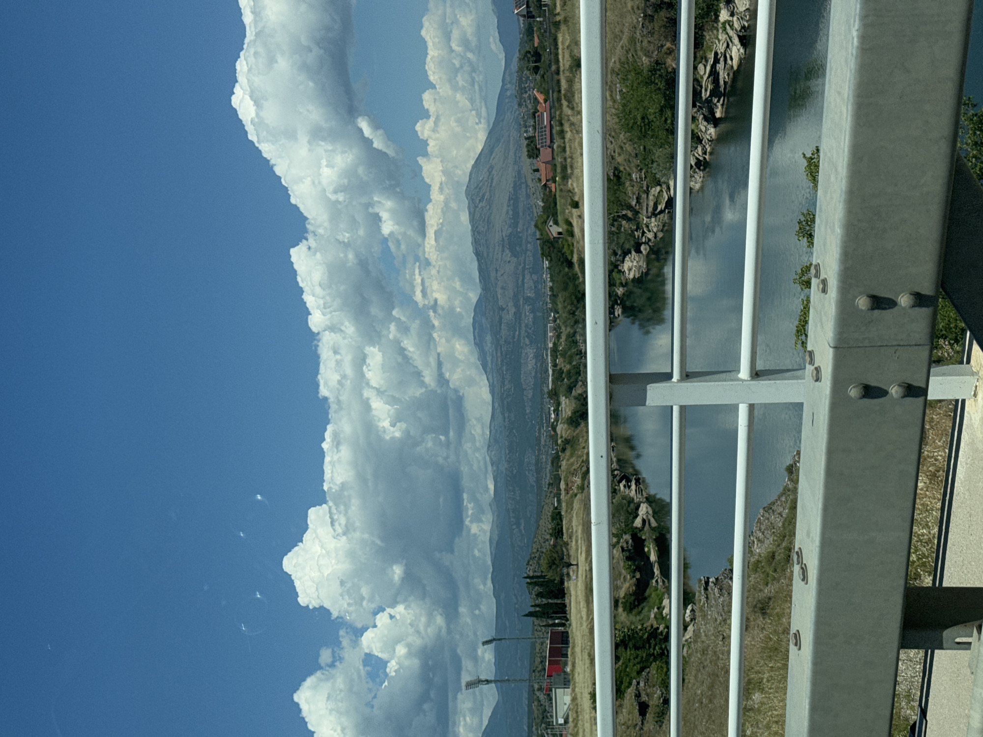 The image depicts a river flowing through a landscape in Podgorica, Montenegro. A bridge railing, constructed from white metal bars and a grey metal beam with visible bolts, dominates the foreground. The river, reflecting the sky, flows through a rocky, vegetated gorge. In the middle ground, dry grass covers rolling hills. Further back, on the left, are two tall stadium lights and a red building. A cluster of cypress trees stands between the lights and the background. In the distance, a range of mountains under a cloudy blue sky forms the horizon. Several buildings with red roofs, some with solar panels, are visible on the hills in the middle to far ground. The scene appears to be a sunny day.
