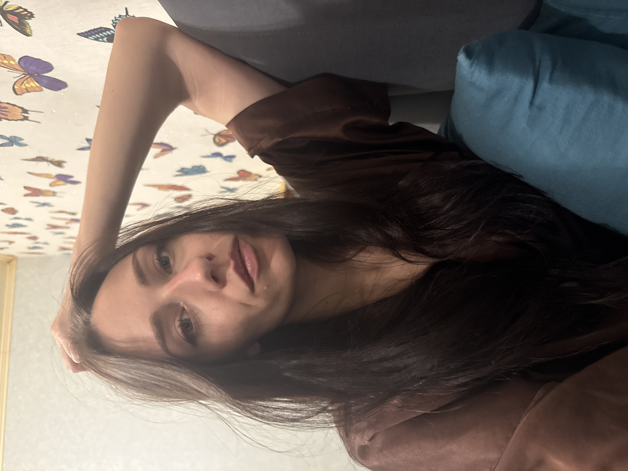 A young woman with long dark hair is depicted in a relaxed, reclined pose, looking directly at the camera. She has fair skin, dark eyebrows, and wears a brown, possibly satin, long-sleeved top or dress. Her lips are a soft pink. Her right arm is bent, resting behind her head or on the surface behind her, while her expression is neutral to slightly pouting, with her eyes engaged with the viewer.
To her right is a light blue/teal pillow, with a dark grey sofa or headboard visible behind it. The wall in the background features numerous colorful butterfly decals in shades of purple, orange, blue, and yellow on a light-colored surface. A partial view of a light-colored frame or trim is on the left side of the wall.
The lighting is soft, indicating an indoor setting, likely a bedroom or living room. No specific time of day or weather can be discerned, and there is no visible text in the image. The location context provided is Saint Petersburg, Russia.
