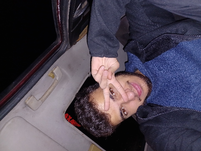 An individual is seen from a slightly elevated angle, possibly inside a car. He is making a hand gesture resembling a peace sign in front of his eye. He wears a blue textured sweater and a dark jacket. The interior of what appears to be a car is visible, including a handle and part of the roof. The background shows a dark space with minimal light.