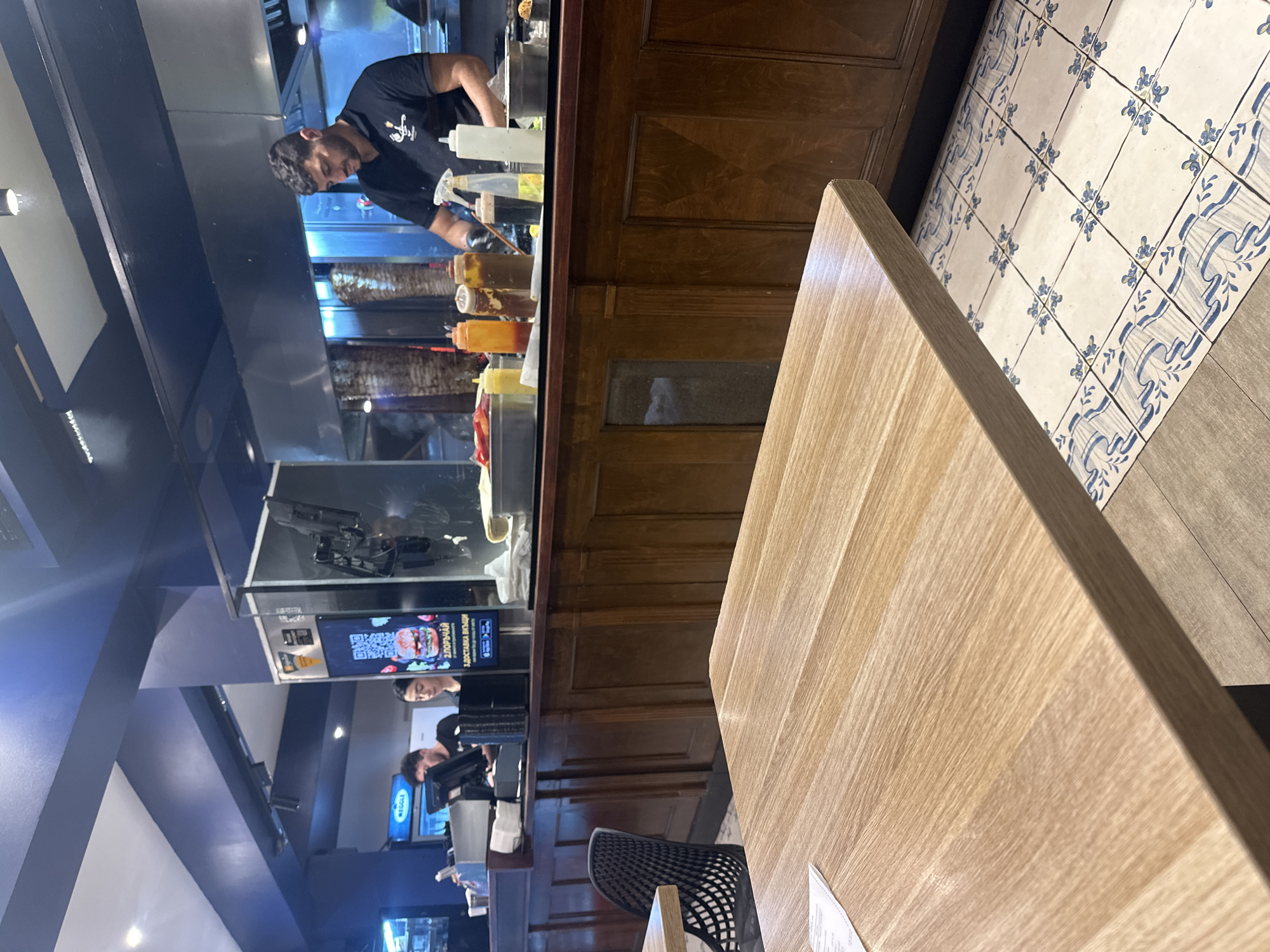 The image shows the interior of a food establishment, possibly a shawarma or kebab restaurant, in Sofia, Bulgaria. A man wearing a black t-shirt is preparing food behind a counter with various sauce containers. Behind the counter, one can see the vertical rotisseries with meat. In front of the counter are a few people working on computers. The floor has tile designs, and a light-colored wooden table dominates the foreground. A modern, mesh-backed chair is placed by the table.