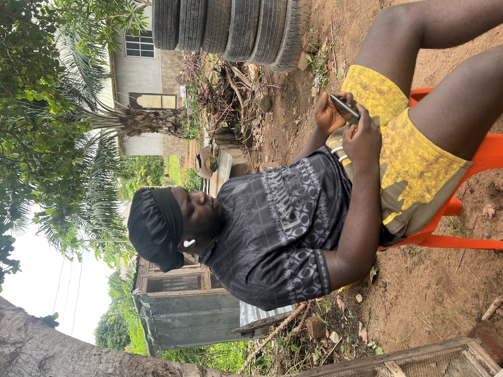 An adult male sits in an orange plastic chair in an outdoor location, appearing to be in Markurdi, Nigeria. He is wearing a black patterned shirt, yellow patterned shorts, and a black head covering. He has an earbud in his left ear and is holding a smartphone in his hands, apparently focused on it. Behind him are stacked tires, a palm tree, and a building. The ground is dirt with scattered vegetation.