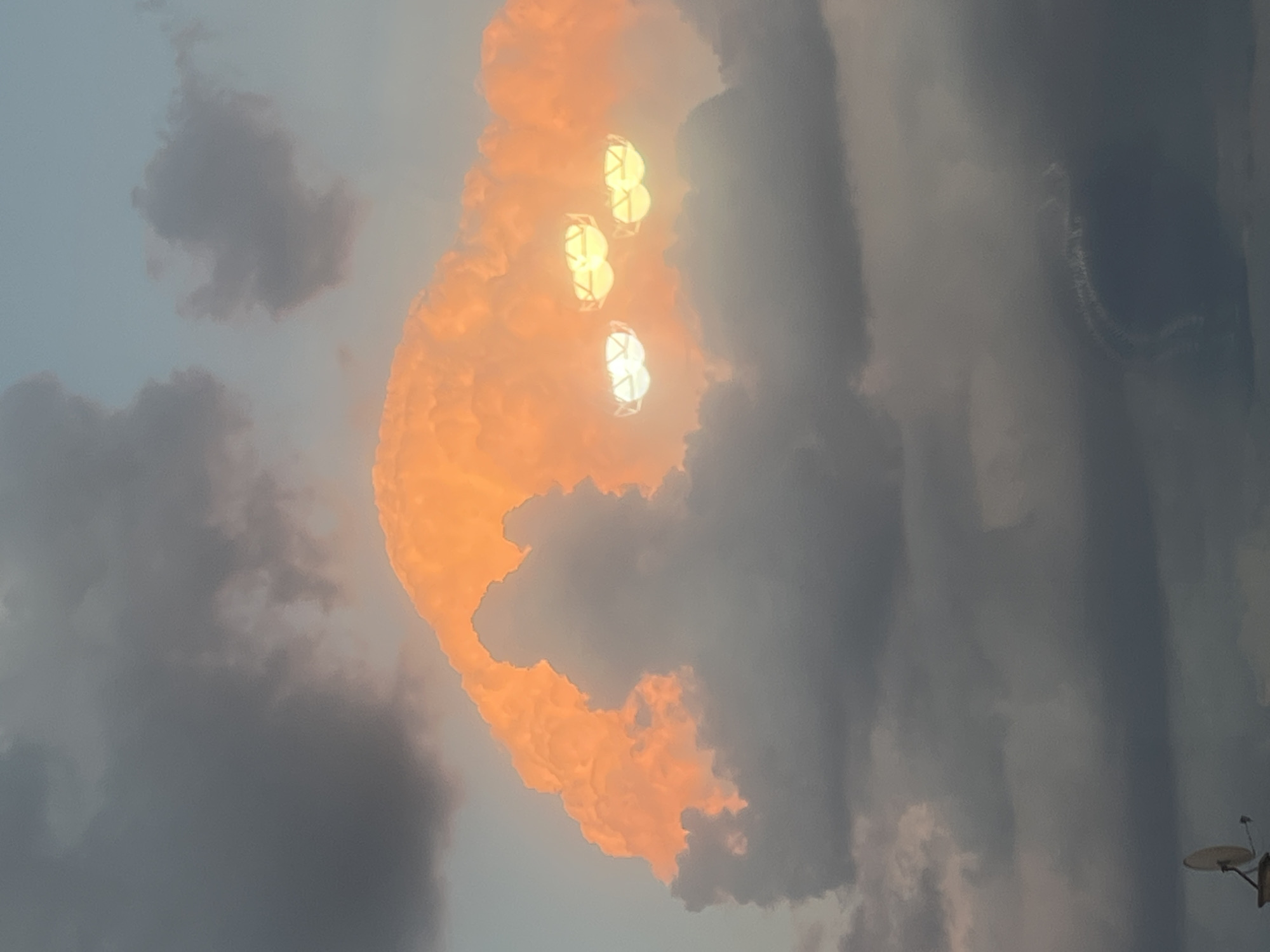 In the evening sky above Cheshnegirovo, Bulgaria, a large, luminous cloud formation is visible, tinged with vibrant orange from the setting sun. Embedded within this cloud are three distinct, glowing orbs that resemble spotlights, casting a bright, diffused light. These orbs are positioned within the cloud, appearing almost as if they are part of its structure or illuminating it from within. Below this prominent formation, darker, more diffused clouds fill the lower portion of the frame. In the bottom left corner of the image, a satellite dish mounted on a structure is partially visible, suggesting a human presence or habitation in the area. The overall scene captures a dramatic interplay of light and cloud during twilight.