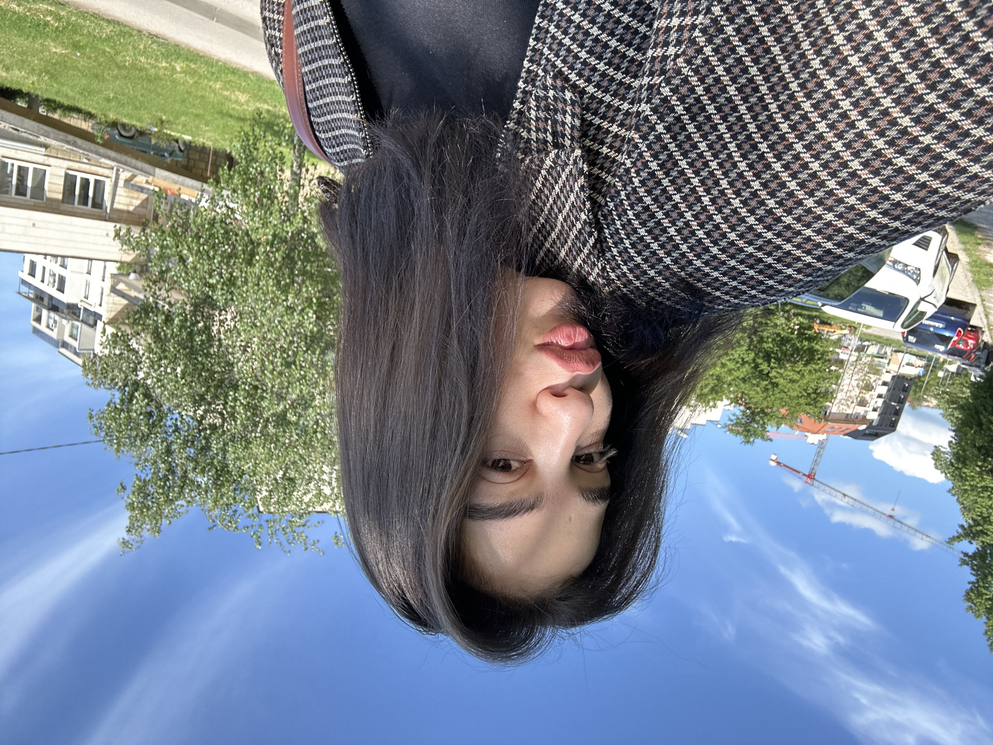 A vertical-format selfie shows a person outdoors. The person is wearing a black top and a brown and black houndstooth blazer. The background features a blue sky with white clouds, green trees, buildings, a construction crane, a street, and parked cars. The location is Sofia, Bulgaria.
