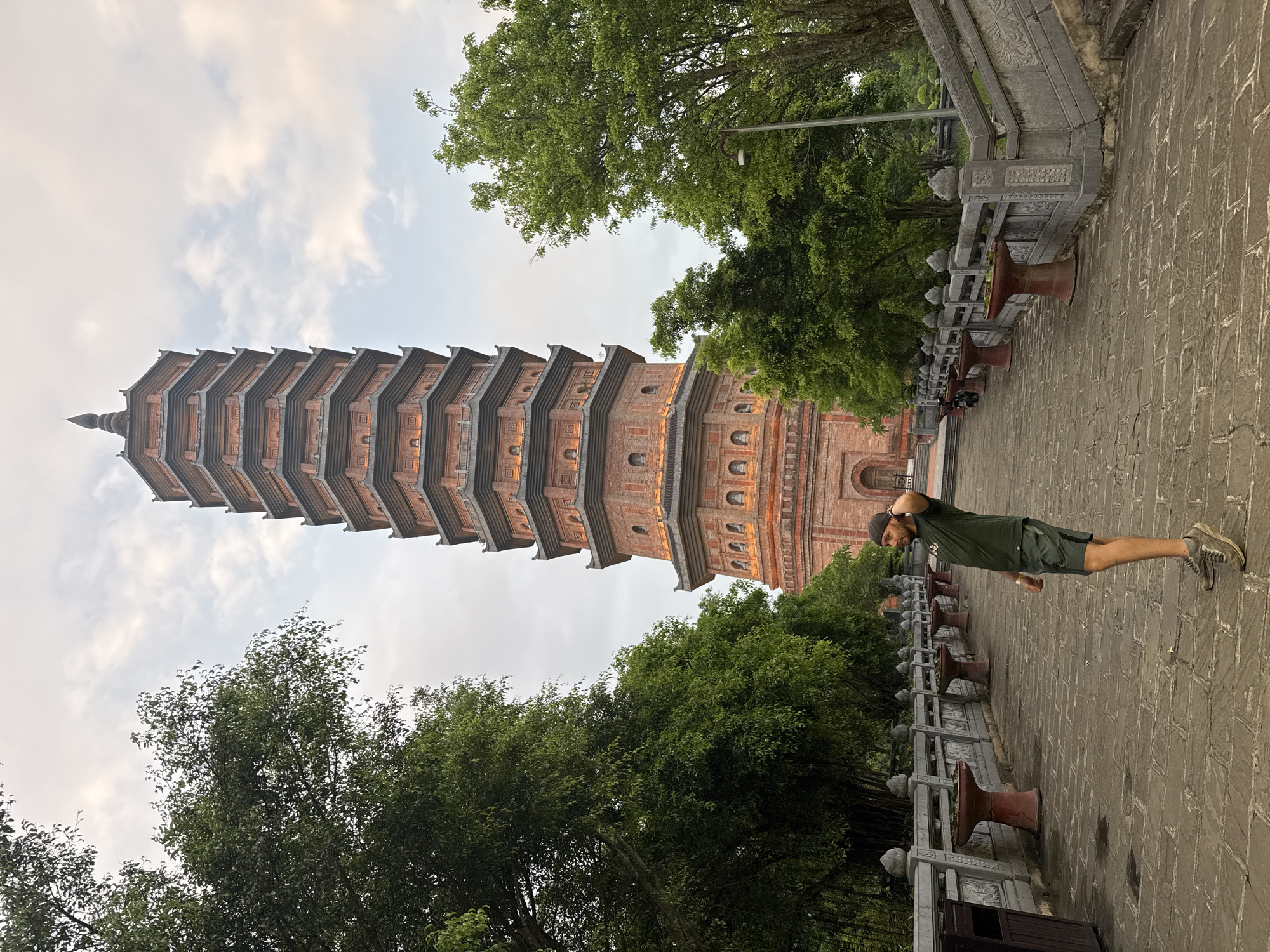 A man wearing a dark green t-shirt and shorts stands in the foreground of an image that features a tall, multi-tiered pagoda in Tây Hoa Lư Ward, Vietnam. The pagoda, constructed of reddish-brown brick with gray roofing, dominates the background. It is adorned with decorative elements and has numerous windows, some illuminated. The man is facing to the right, with his left hand on the back of his head and his right arm slightly bent. He is standing on a paved walkway made of stone slabs.
The walkway is flanked on the left by a stone railing with decorative carvings and several urn-like planters. On the right, a set of stone stairs leads upward. Lush green trees with dense foliage are visible on both sides of the walkway and behind the pagoda. The sky above is overcast with scattered clouds. The overall scene suggests a historical or religious site within a natural setting.