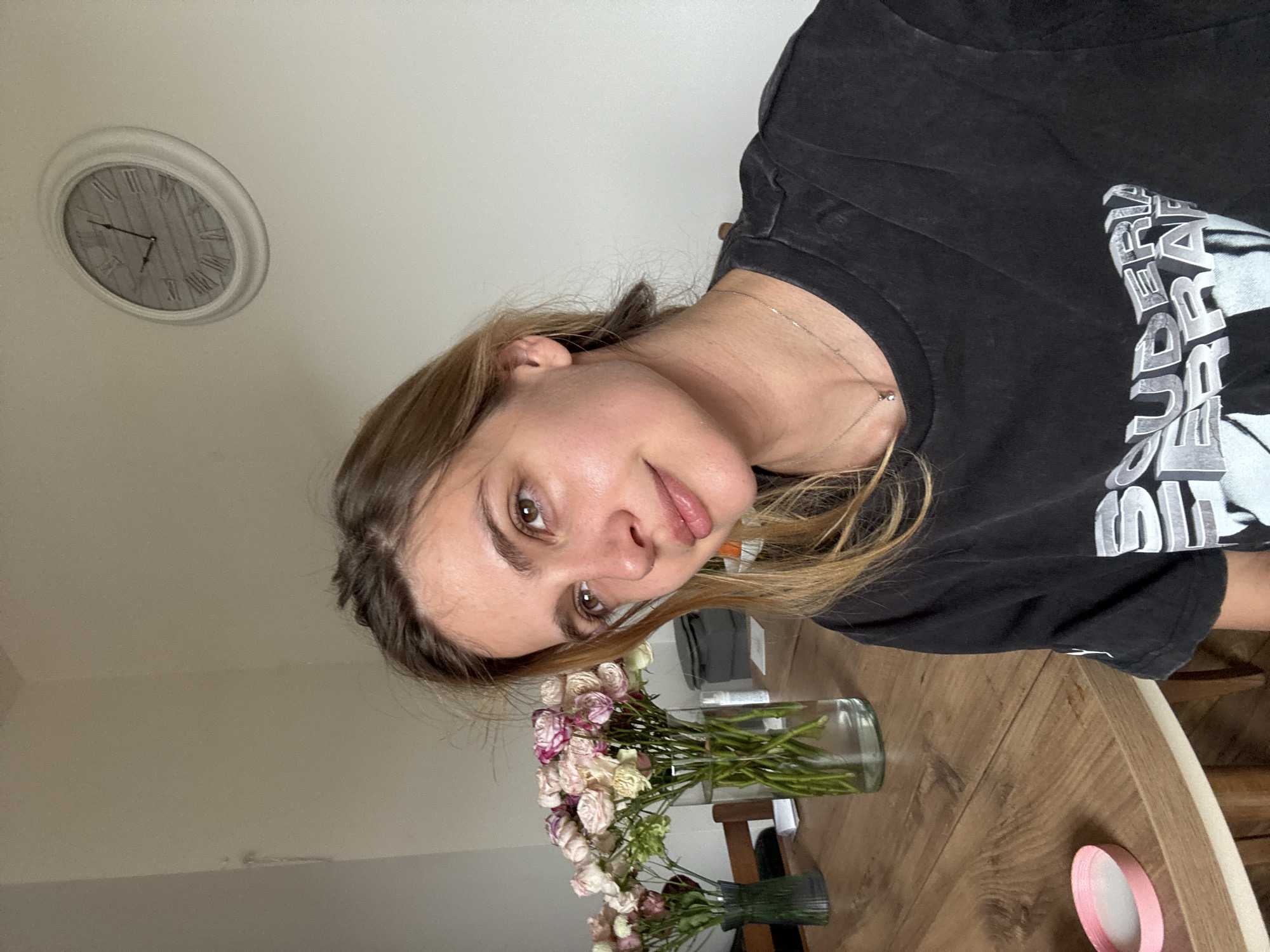 A person is visible in the image. The person has brown hair and is wearing a black T-shirt. The person is positioned near a wooden table on which flowers and a vase are present. Also on the table is a pink circular object. On the wall is a clock. The location is Unknown, Unknown.