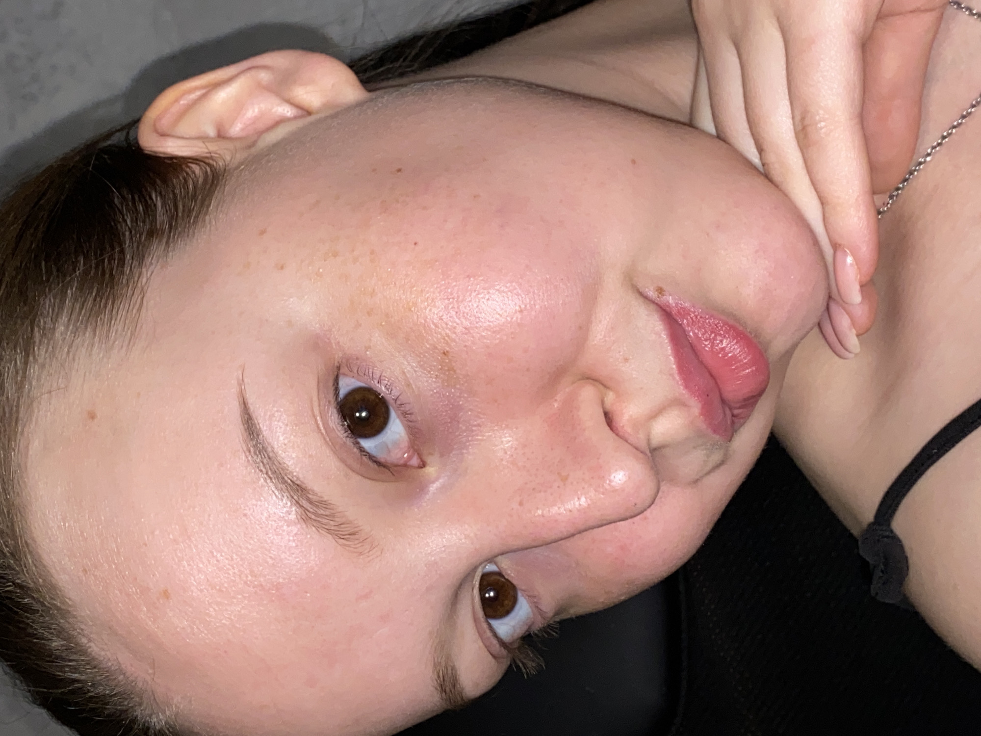 A close-up, tilted perspective photo shows a fair-skinned person with brown eyes and light brown hair. Freckles are visible on their face. The person's lips are slightly puckered. A hand is touching the person's neck, where a thin silver necklace is visible. A black strap is also visible on the person's skin. The background is indistinct. There is no information available to determine the location of the scene as Chaikovsky, Russia.