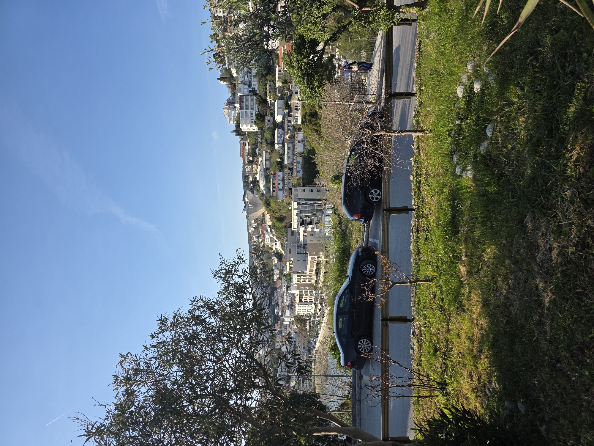 The image shows a view of the city of Ulcinj, Montenegro from an elevated position. In the foreground, there is a road with two dark colored cars parked along the side. The road is bordered by a green, grassy area. Beyond the road and cars, the city stretches out towards the sea. Buildings of various sizes and styles are densely packed together. The sky is clear and blue. There are trees in the image frame. Two people are sitting on a bench in the background of the image.