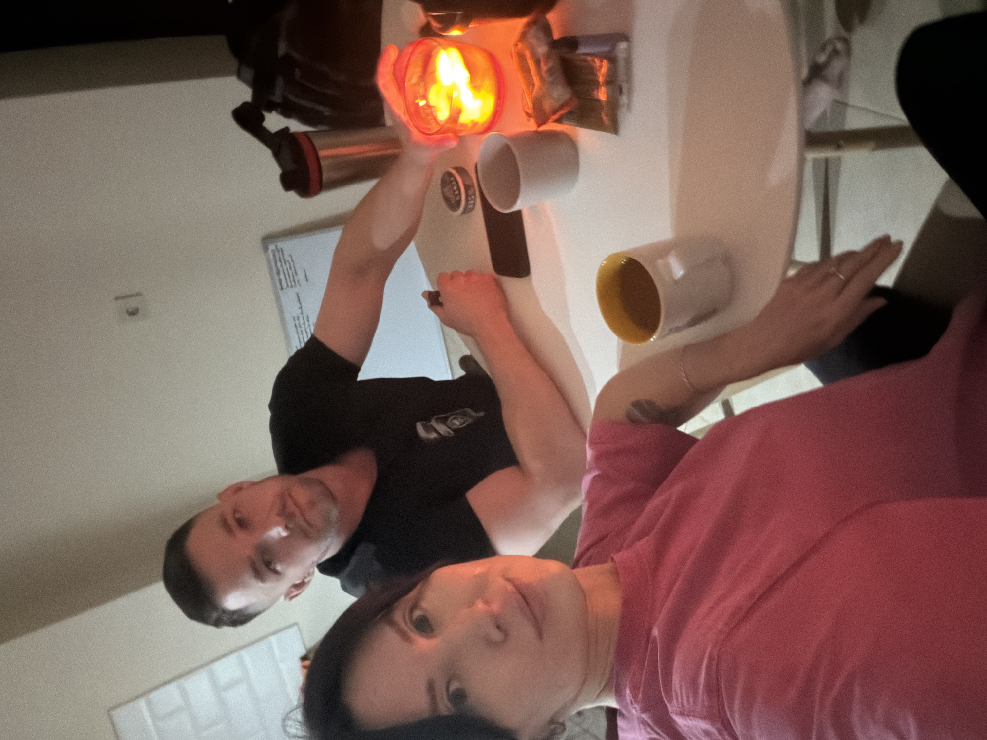 A selfie captured indoors, presumably in the city of Perm, Russia, features a man and a woman seated at a white table. The man, on the left, wears a black t-shirt. The woman, in the foreground, wears a pink shirt. On the table are two mugs (one white and one yellow), a metallic container, an orange, possibly flammable, object, a mobile phone, and other unidentifiable items. The overall lighting in the scene is warm.