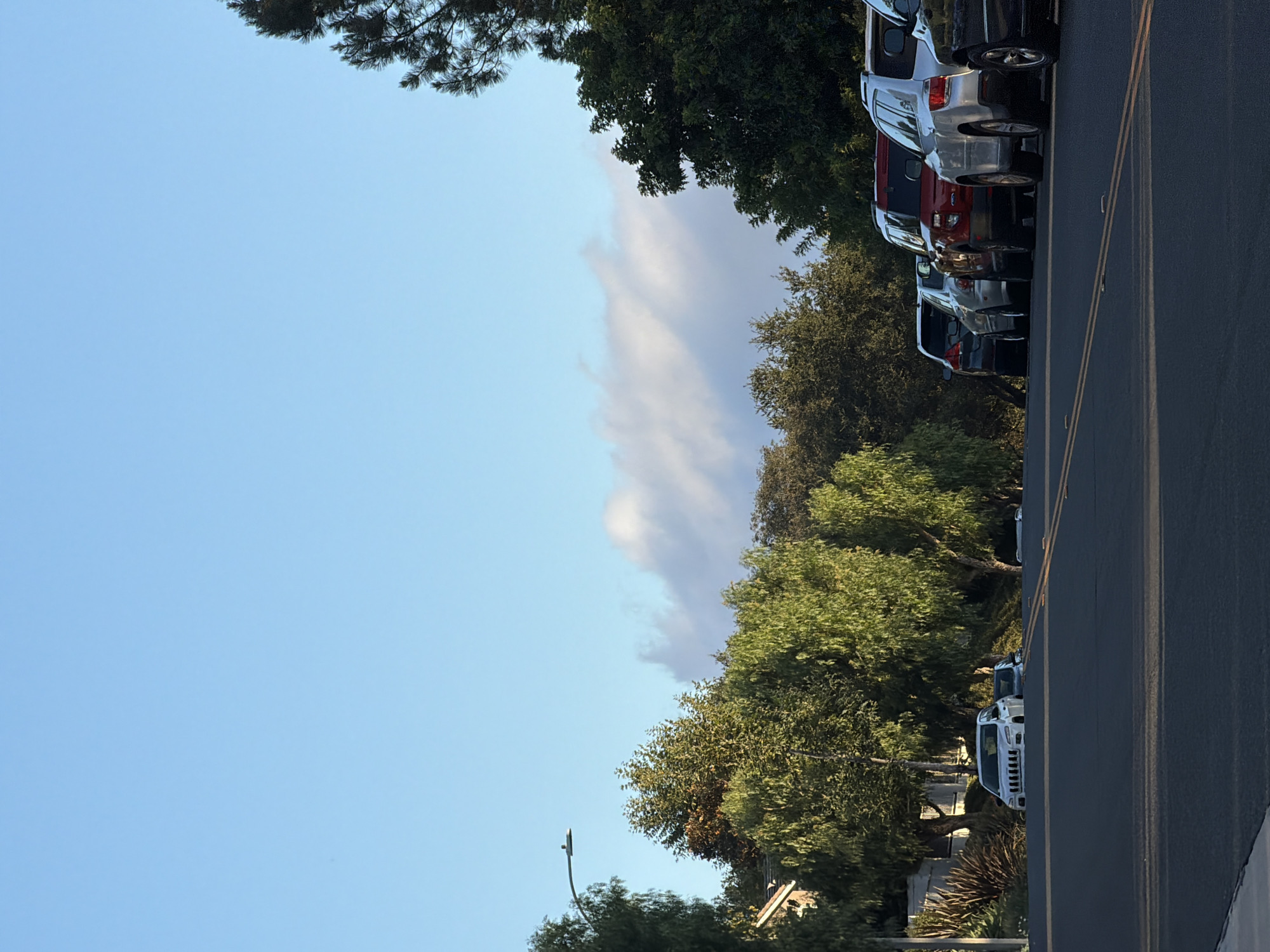 The image depicts a street scene in Aliso Viejo, California, with parked cars lining the right side of the road. The perspective is from the left side of the road, looking towards the parked cars and the sky. The sky is a clear, pale blue, with a thin, wispy cloud formation stretching across the upper half of the image. Sunlight suggests it is daytime, likely late afternoon or early morning, given the softness of the light and the long shadows cast by the trees.
A line of various vehicles is parked along the curb, including SUVs and a white truck. The cars are facing away from the camera, suggesting they are parked along a one-way street or in a parking lot adjacent to the road. Lush green trees and foliage are visible behind the parked cars, with some branches extending into the upper portion of the frame. The road itself is paved and marked with yellow lines. A streetlight is visible in the lower left quadrant of the image, emerging from the trees. The overall atmosphere is peaceful and suburban. No people are visible in the image.