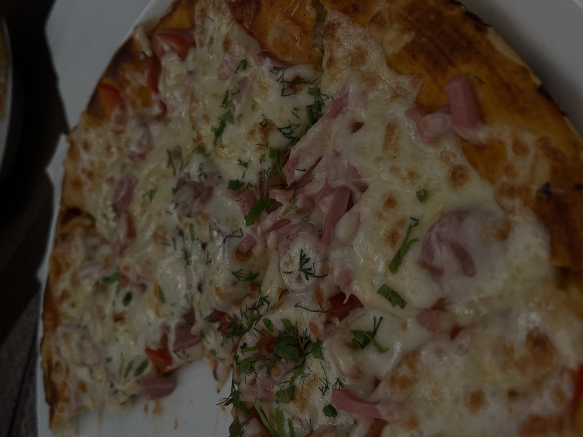 A close-up, high-angle shot captures a partially consumed pizza resting on a white, round plate in what is identified as the city of Novopokrovskaya, Russia. The pizza features a golden-brown crust and is generously covered with melted white cheese, displaying slight browning in various spots. Distributed across the cheese are numerous pieces of light pink, processed meat, consistent with ham or sausage, cut into irregular strips and chunks. Red bell pepper segments, varying in size, are also visible among the other toppings. Fresh green herbs, primarily dill and possibly parsley, are sparsely sprinkled over the entire surface. One slice of the pizza has been removed from the left side, leaving an empty space on the plate. The background is blurred, showing faint outlines of other objects, suggesting a dining setting.