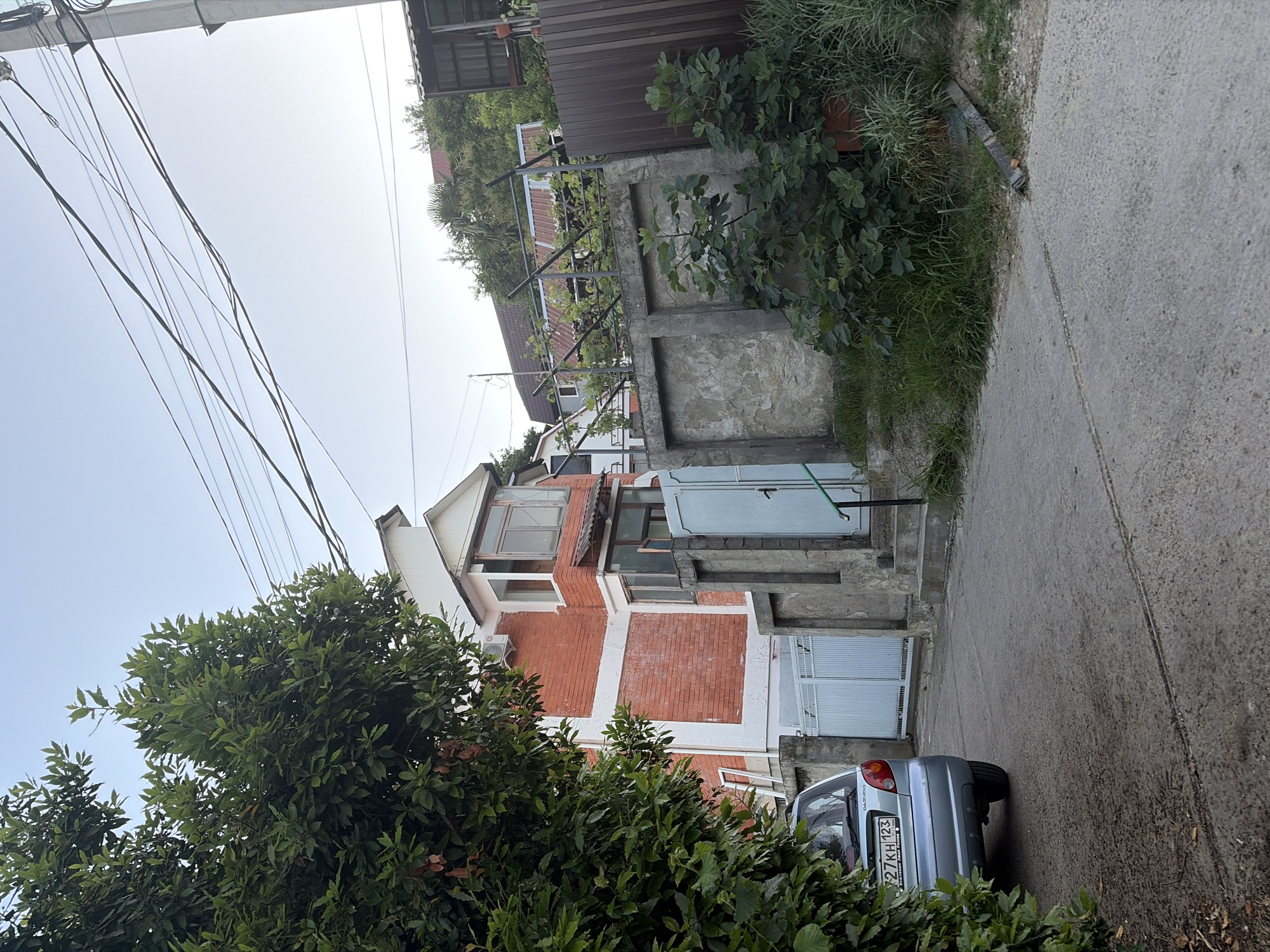 A view from a low angle shows a street in Sochi, Russia, with a car parked on the left side. The car is light blue and has a license plate. To the right, there is a brick building with white trim, a light blue gate, and a concrete fence. In the background, there are trees and other buildings. The sky is clear and blue. There are electrical wires strung across the sky. The overall scene appears to be a residential area.