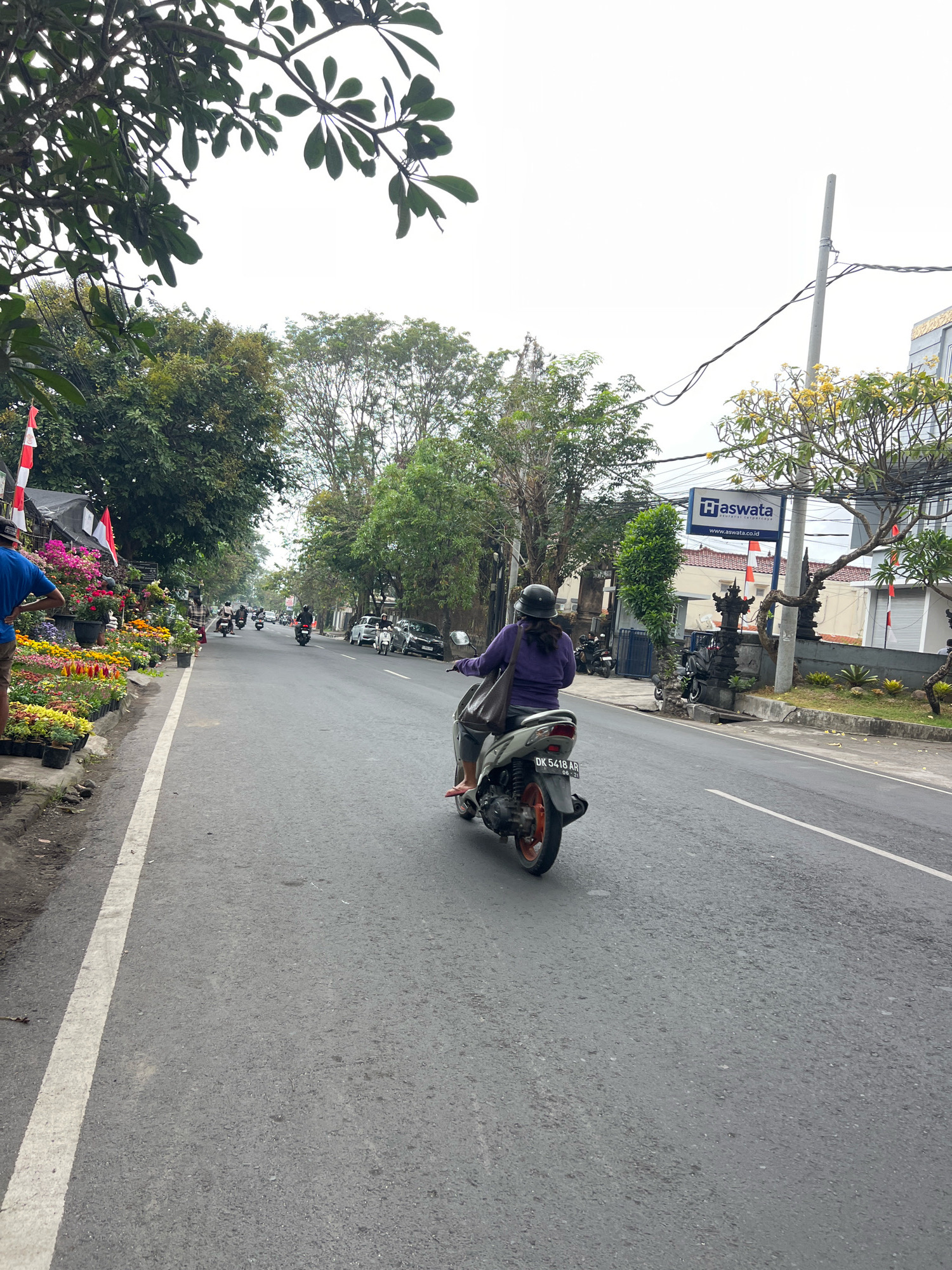 The image depicts a street scene in Denpasar, Indonesia. A person wearing a purple top and a helmet is riding a scooter on the right side of the road, moving away from the camera. The scooter has a license plate that reads "DK 5418 AR". On the left side of the road, there is a flower stall with a variety of colorful potted plants, including pink and yellow flowers. Several Indonesian flags are visible on poles above the flower stall. Further down the street, there are other scooters and cars. A building with a sign that says "ASWATA" is visible on the right side of the road, indicating it is an insurance company. The sky is overcast, and the street is paved with asphalt. Trees line both sides of the road, and there are power lines overhead. The overall impression is a busy street with ongoing traffic and local commerce.
