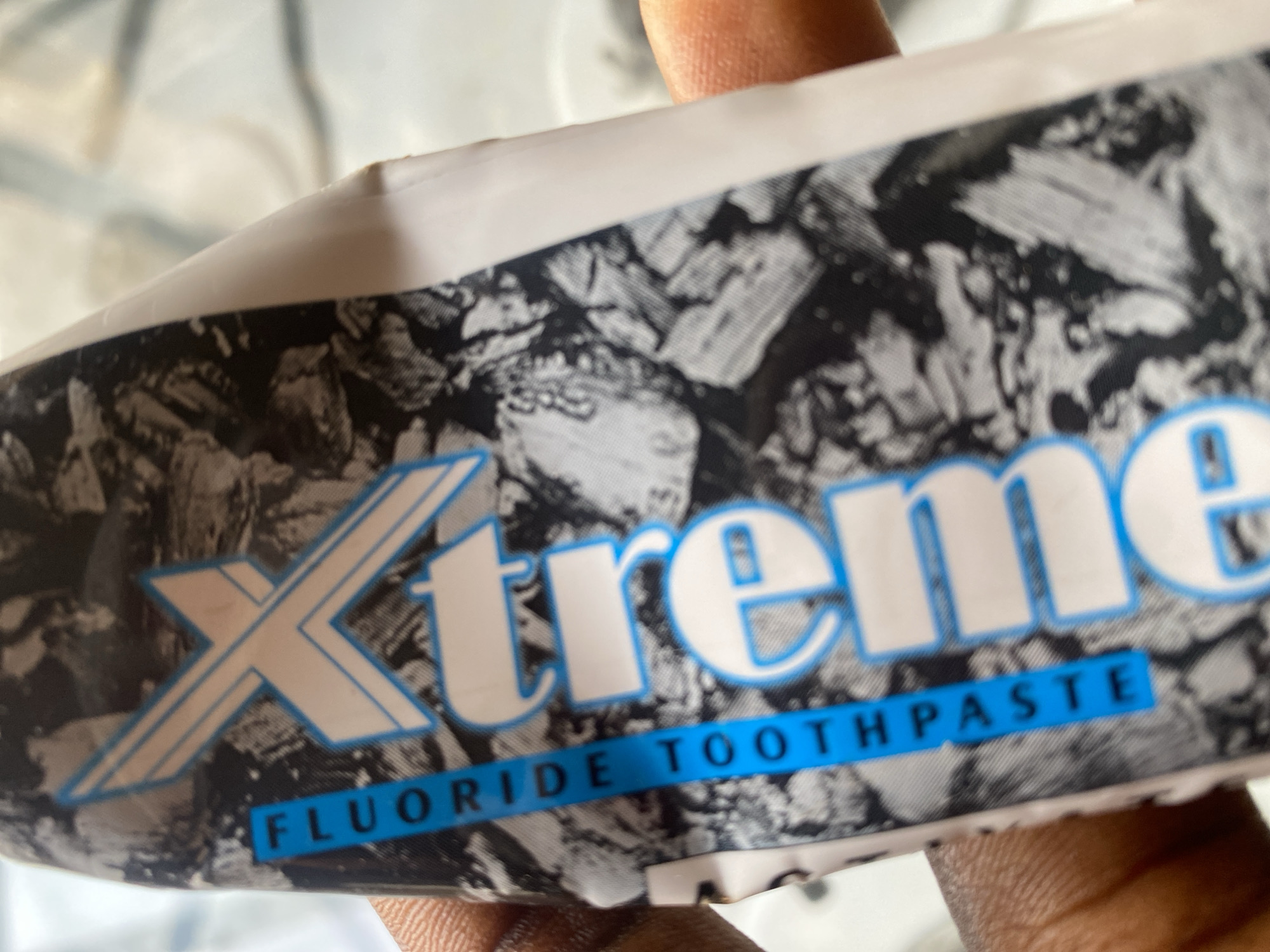 A hand is holding a tube of "Xtreme Fluoride Toothpaste." The tube has a black and white, abstract, rocky pattern as its background. The brand name "Xtreme" is in large, white, capitalized letters with a blue outline. Below the brand name, on a blue stripe, the words "Fluoride Toothpaste" are written in white, capitalized letters. The image appears to be taken indoors, possibly in a home or a shop, given the lighting. The hand holding the toothpaste suggests it is being presented or examined. The overall scene is a close-up of the product, emphasizing the branding and product type. The location context of Jauro Yinu, Nigeria, is provided but not visually evident in the image itself.