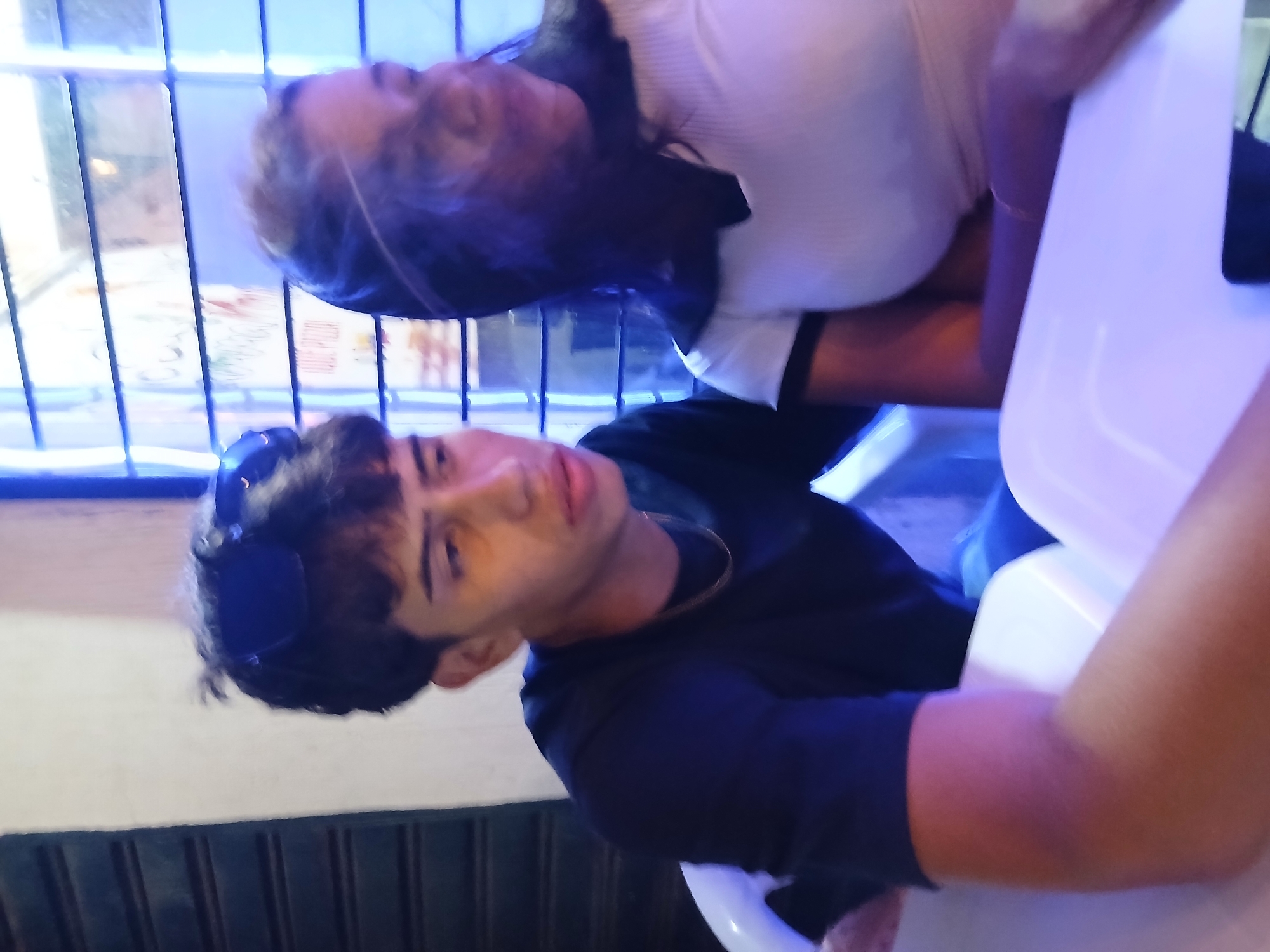The image, taken in Juan Griego, Venezuela, depicts two young individuals in an indoor or covered outdoor setting, possibly a cafe or restaurant, illuminated by a strong blue light suggesting an evening or nighttime scene. The photograph is oriented at an angle, showing the subjects as if leaning.
In the foreground, a young male is seated, looking towards the camera. He has short, dark, wavy hair with sunglasses pushed up onto his head. He is wearing a dark, possibly black, long-sleeved shirt and a thin gold chain around his neck. His expression is neutral. His left arm is visible, resting on a white plastic surface, likely an armrest or part of a chair.
Behind him, a young female is also seated, positioned slightly above and behind the male. Her head is tilted back, and her face is partially obscured by her dark hair. She appears to be wearing a light-colored (possibly white or off-white) top with a ribbed texture. Part of her arm, covered by a dark sleeve, is visible, and she is leaning against a white surface, consistent with the plastic seating in the venue.
The background features vertical metallic bars, possibly part of a window, railing, or decorative screen, which brightly reflect the blue ambient light. White plastic furniture, including chairs and potentially a table, is visible throughout the scene. There is no visible text in the image. The overall atmosphere appears casual and relaxed.