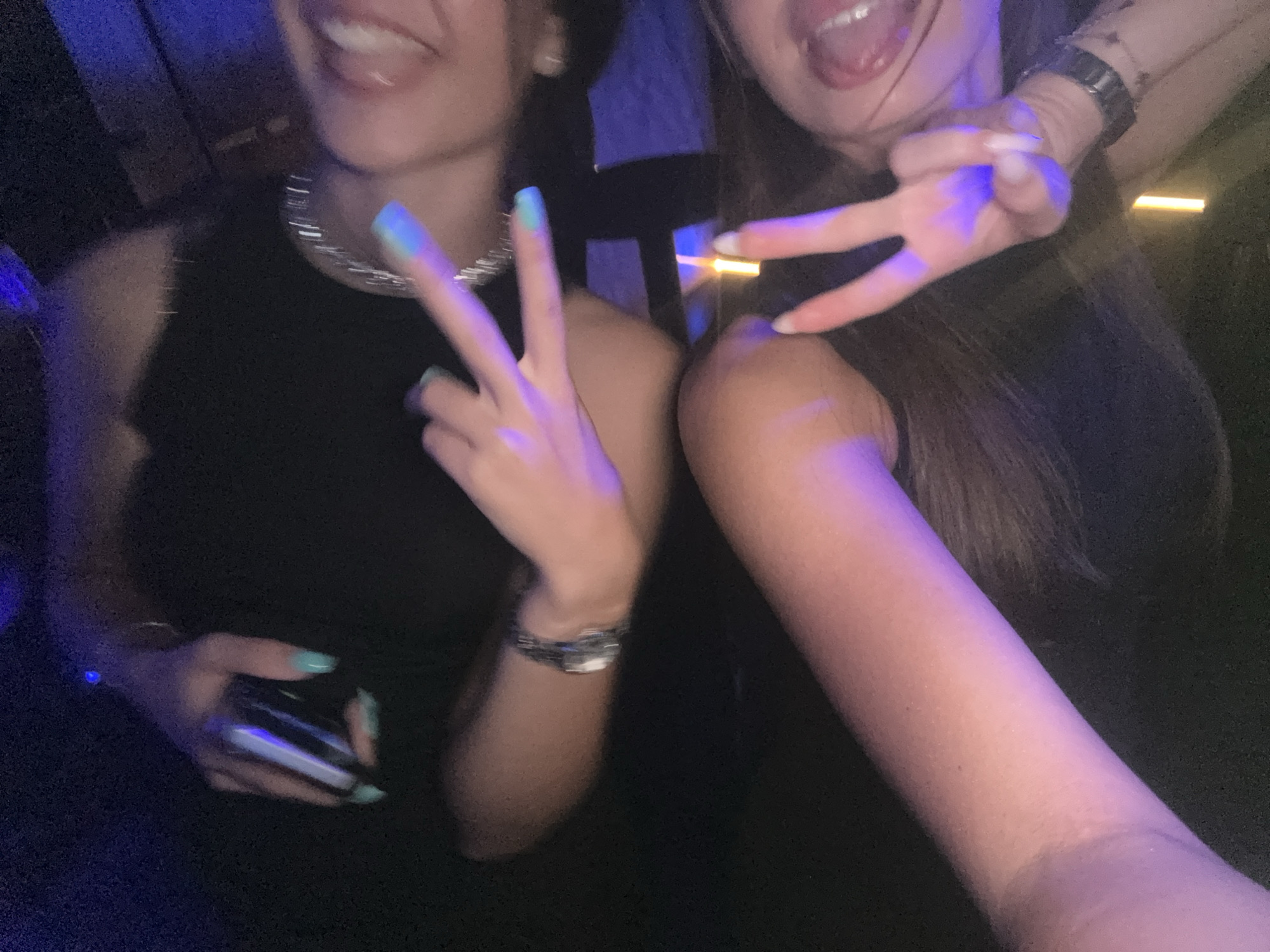 The image shows two individuals, their faces partially visible and illuminated by blue and purple light. One person on the left is wearing a black top and a silver chain necklace, with light blue nail polish. They are holding a dark object that appears to be a phone. The other person on the right, also in dark clothing, is wearing a watch on their left wrist and has their tongue sticking out slightly, making a peace sign with their right hand. The environment appears to be indoors, possibly a club or dimly lit venue. The scene does not provide any specific information to determine the location within Sozopol, Bulgaria.
