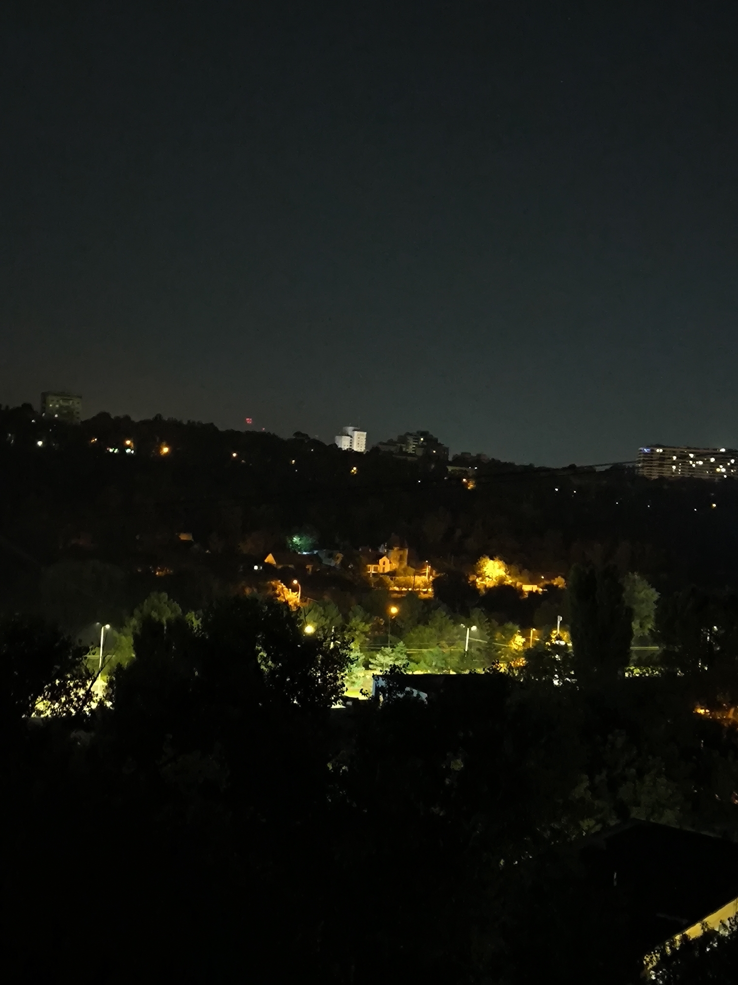 This is a nighttime, elevated shot of a semi-urban landscape in Chișinău, Moldova. The sky is dark with no visible stars or moon. In the foreground, dense foliage creates a dark, shadowy area, obscuring much of the lower part of the image. Further back, the rolling hills are dotted with lights from buildings and streetlamps, indicating the presence of a populated area.
Several buildings are visible on the distant hills. To the left, a multi-story building with illuminated windows stands on a ridge. In the center, a cluster of lights suggests a residential area or a small town. Further to the right, a large, modern building with many illuminated windows is prominent on the horizon. There are also scattered streetlights casting a yellowish glow on the landscape. The overall impression is of a quiet, sleeping city seen from a distance at night.