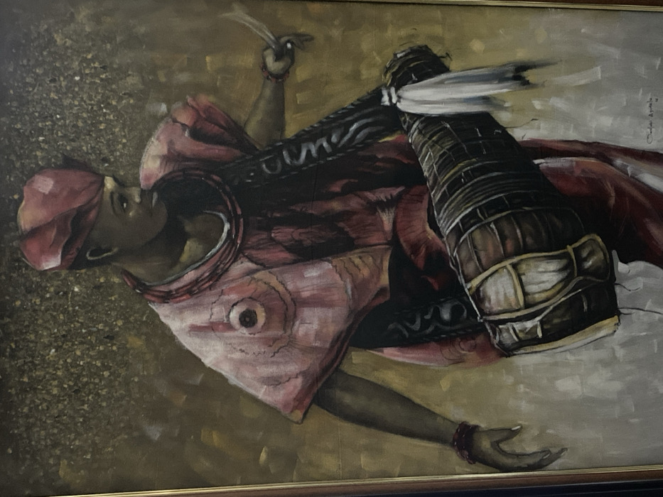 This image is a photograph of a framed oil painting depicting a single individual, likely male, positioned slightly off-center and angled to the right. The subject wears a red traditional cap (fila) and a loose, reddish-pink tunic adorned with circular patterns and darker intricate designs. A dark, patterned strap crosses the subject's chest, securing a brown elongated talking drum (gangan) that rests across the lower body. The drum features white wrappings and decorative tassels. The individual holds two thin, curved drumsticks in the raised right hand and wears red wrist bracelets on both arms. The left hand is slightly open and lowered. The subject's dark skin tone is visible, and the gaze is directed towards the right. The background is an abstract, textured field of shimmering gold, brown, and ochre tones with speckled highlights. The painting is enclosed within a dark frame featuring a thin gold inner border. A signature, partially visible as "Chuks Okonkwo" or similar, is located in the bottom right corner of the canvas. This painting is located in Ikate, Nigeria.