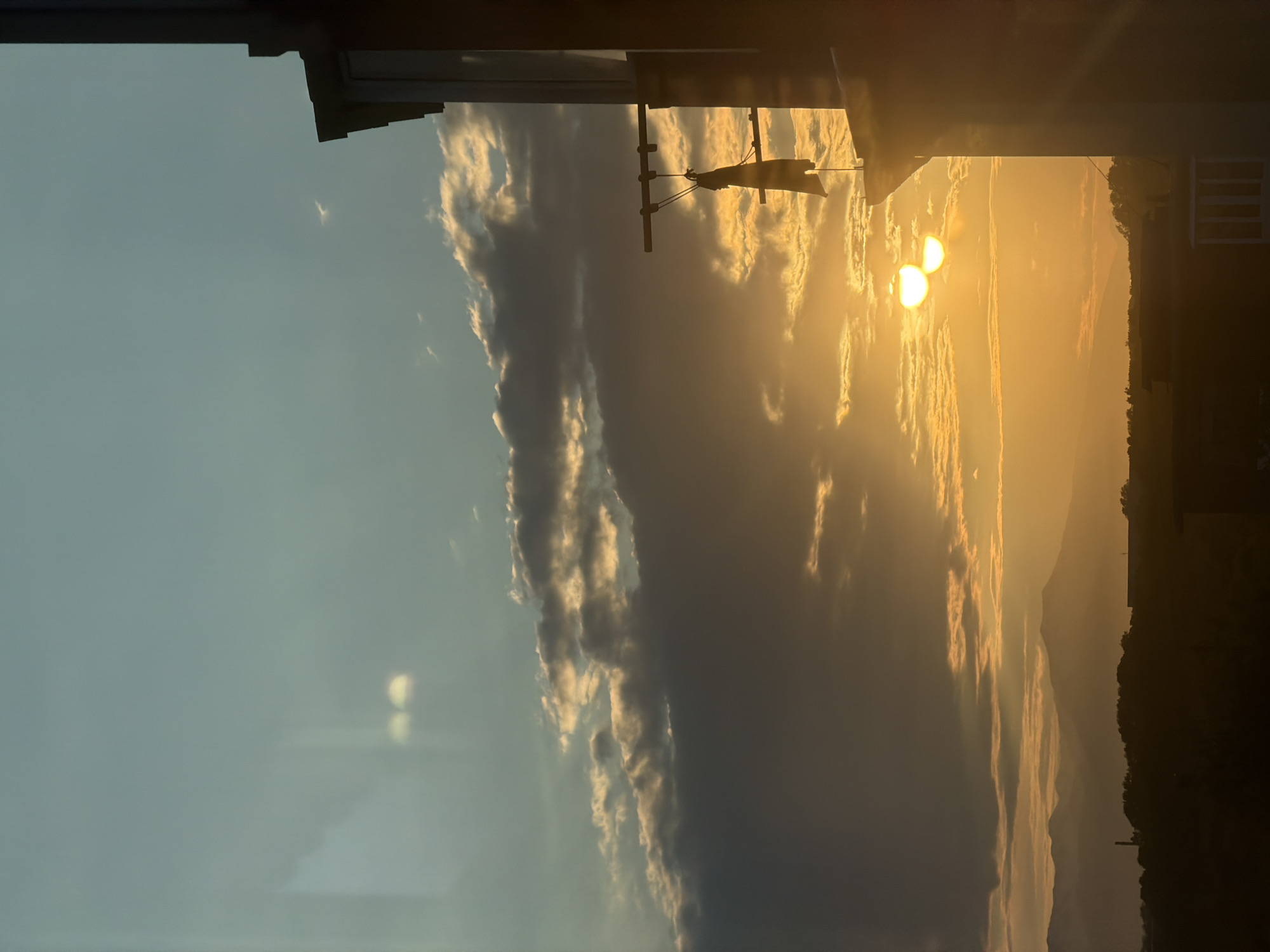 The image captures a sunset or sunrise over a mountainous landscape, with the sun appearing as two bright orbs partially obscured by clouds. The sky transitions from a clear, light blue on the left to a warm, golden hue on the right, illuminated by the setting or rising sun. Wispy clouds are scattered across the sky, catching the sunlight and creating dramatic contrasts between light and shadow.
In the foreground, the silhouette of a building or structure is visible, with dark edges and some indistinct details. A horizontal bar with strings attached, possibly a clothesline, is also silhouetted against the bright sky. In the distance, rolling hills or mountains are visible, also appearing in silhouette against the vibrant sky. The overall scene evokes a sense of tranquility and the beauty of nature's display. The time of day is clearly indicated by the golden light and the position of the sun. The weather appears clear with some cloud cover. There is no visible text in the image. The location context is Petropavlovsk-Kamchatsky, Russia.